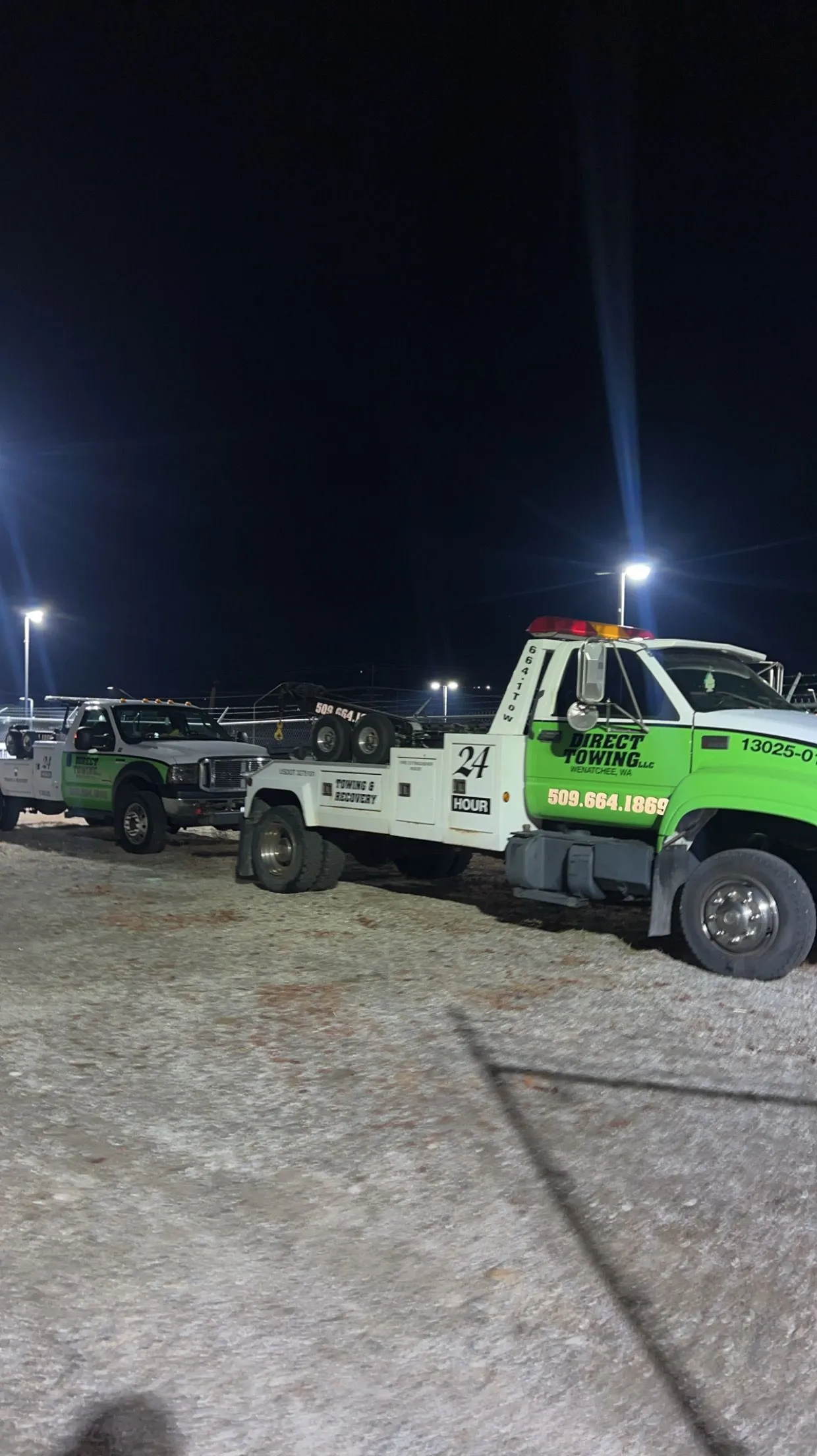 Two Direct Towing recovery trucks parked under bright lights during nighttime operations, showcasing heavy-duty towing capabilities, emergency roadside assistance, vehicle recovery services, and professional fleet readiness.