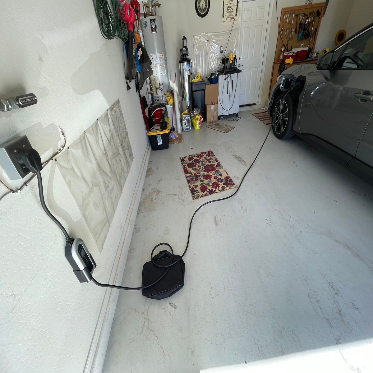 A residential garage features wall-mounted electric vehicle charging equipment connected to a parked gray car, with charging cable extended across the floor near storage tools, water heater, and cabinets nearby.
