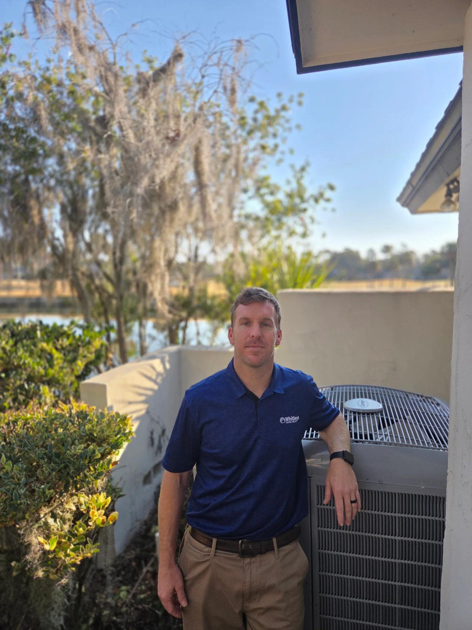 A man in a blue polo shirt with a company logo and tan pants stands next to an outdoor air condition 