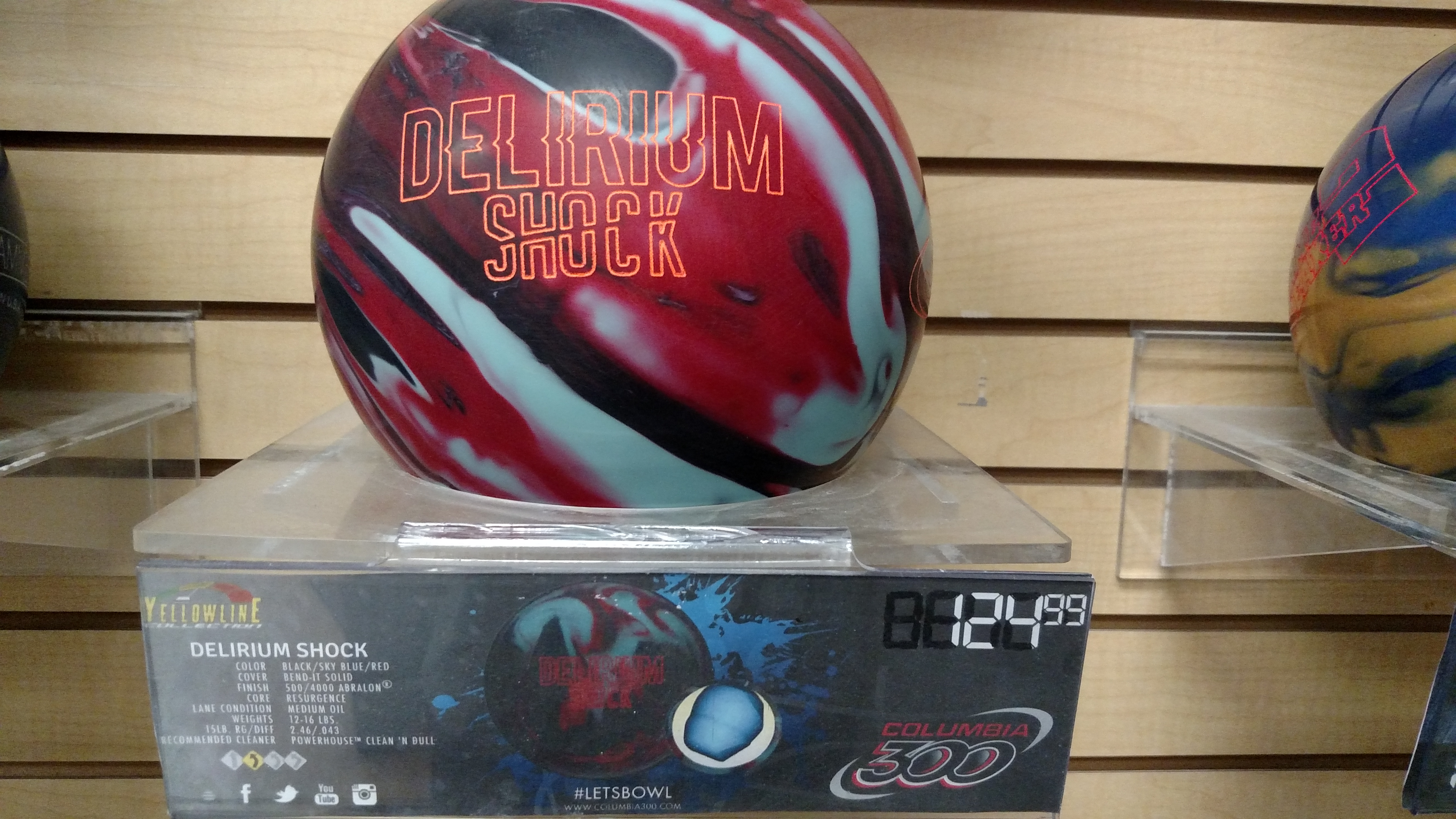 A close-up shot of a bowling ball with the "DELIRIUM SHOCK" design, displayed on a clear stand in front of a wooden slat wall. The ball features a swirl pattern of red, black, and light blue. Below the ball is a display stand with product information and branding for Columbia 300. The price "124.99" is also visible.