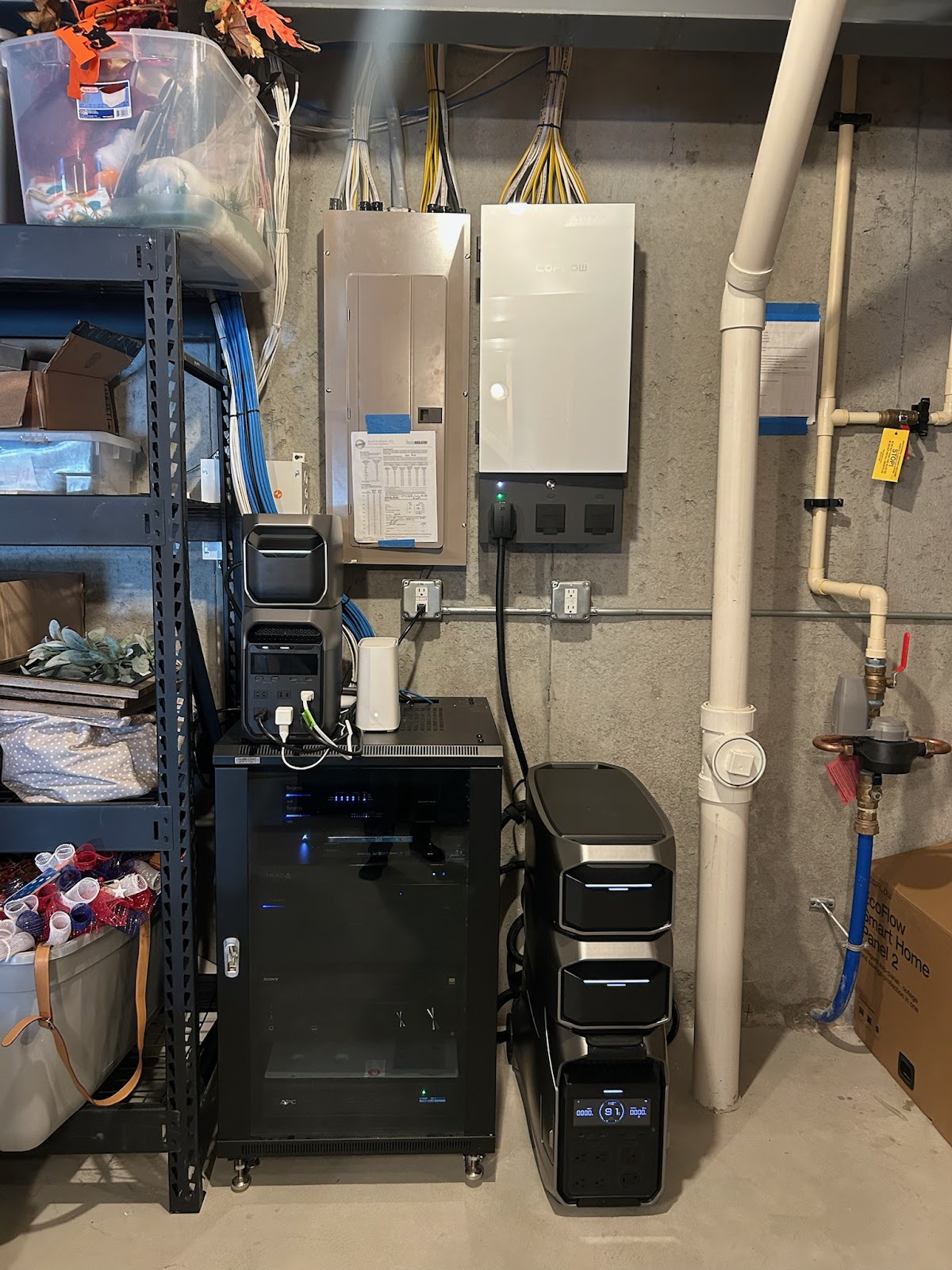 A collection of electronic equipment, including a large white inverter, a gray electrical panel, and several portable power stations, are mounted on or placed near a concrete wall in what appears to be a utility room or basement. The equipment is organized and wired, with cables neatly bundled. Additional items like storage bins and shelving are visible in the background. A water pipe system is also present on the right side of the image.