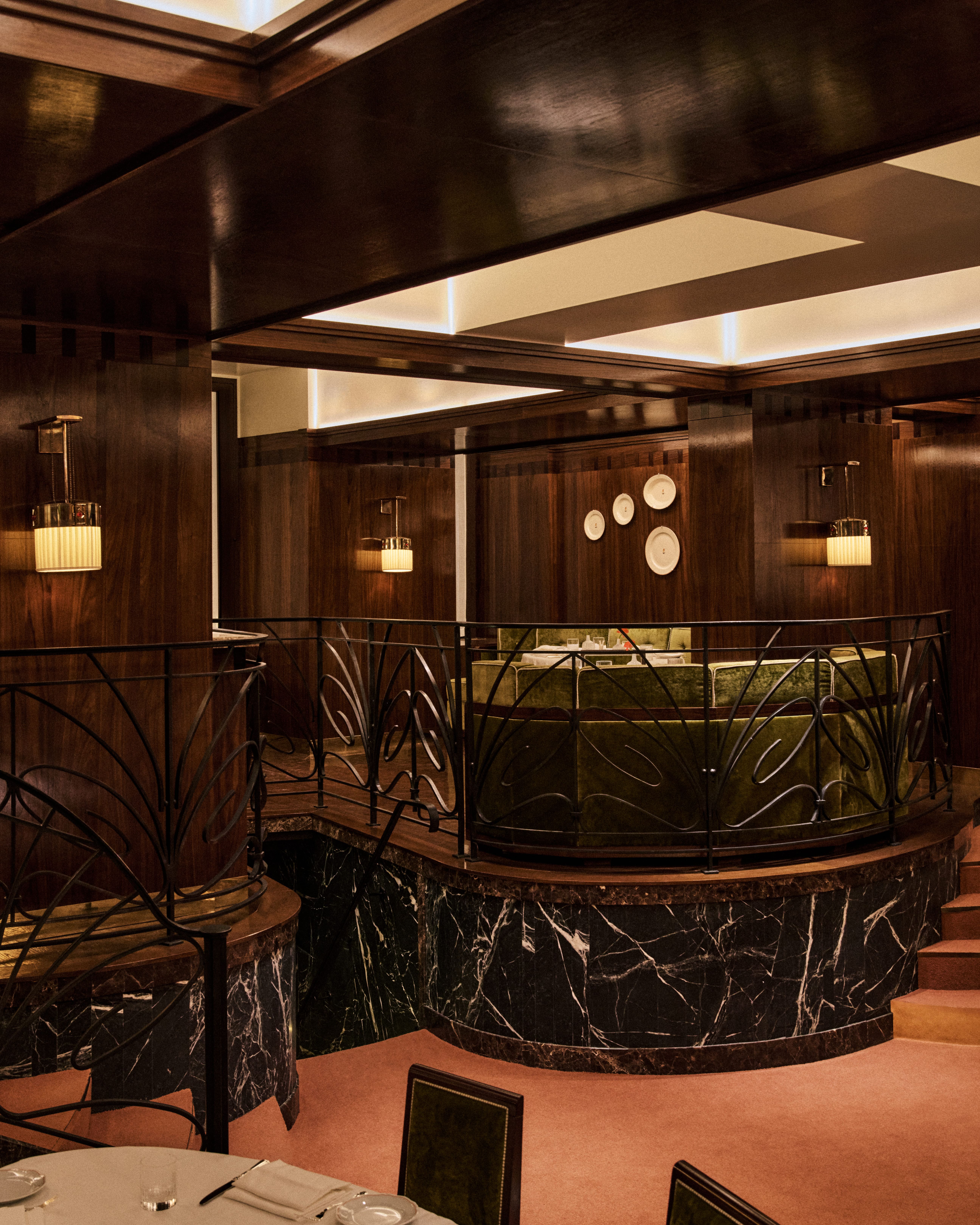 Art Deco–inspired dining mezzanine with curved green velvet banquettes, black marble base, and ornate wrought-iron railings, set against rich wood paneling and softly lit coffered ceilings.