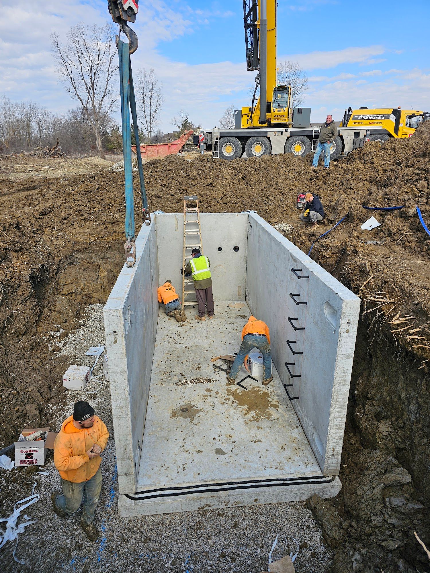 Construction crew installs a large precast concrete utility vault into an excavated trench using a crane. Workers align the structure on a gravel base while preparing underground infrastructure connections.