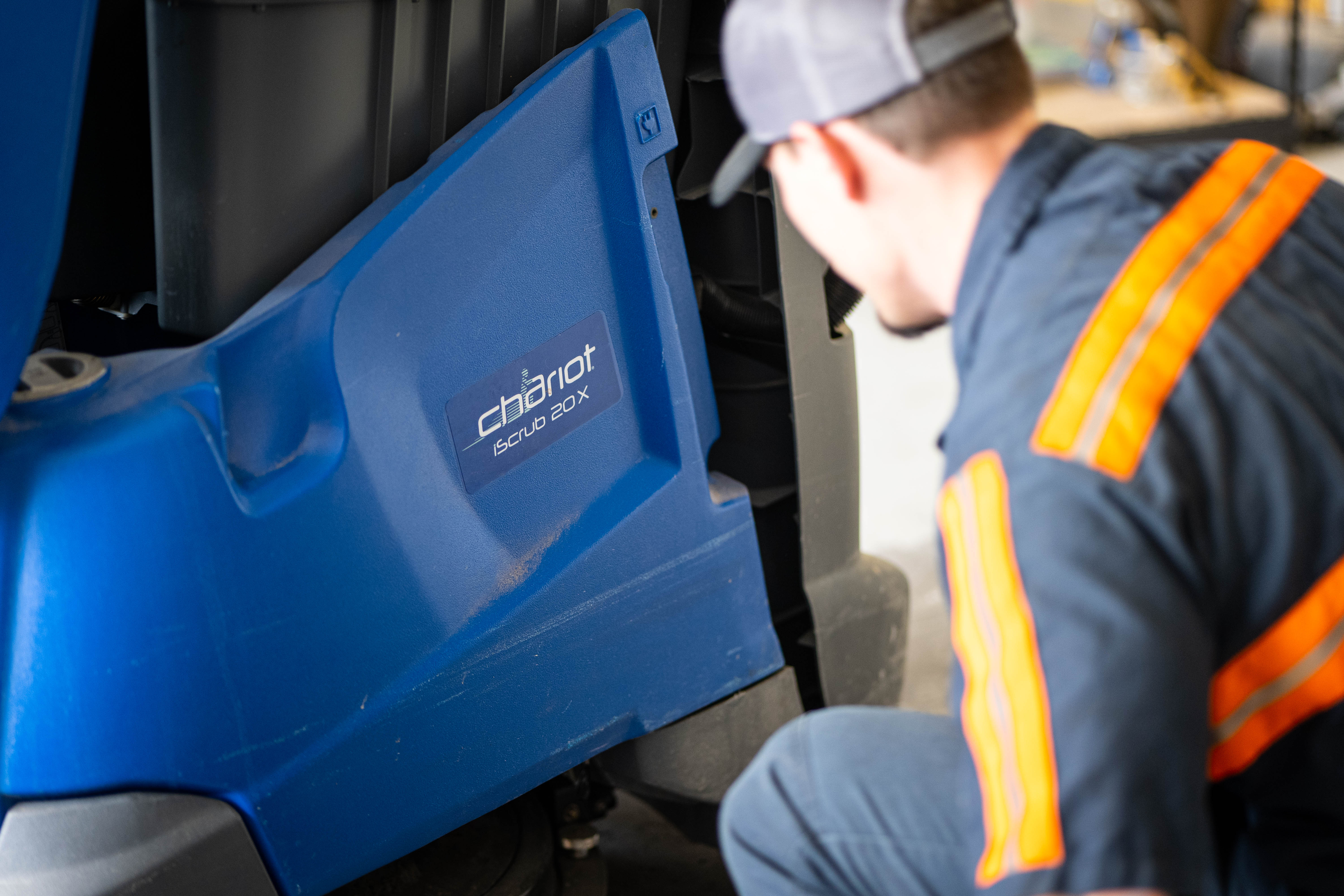 A close-up, high-angle profile shot of a WaySolutions service technician inspecting the lower structural housing and mechanical assembly of a bright blue Kärcher Windsor Chariot iScrub 20X stand-on floor scrubber. The technician is wearing a branded gray work cap and a navy service uniform with bright orange safety stripes.