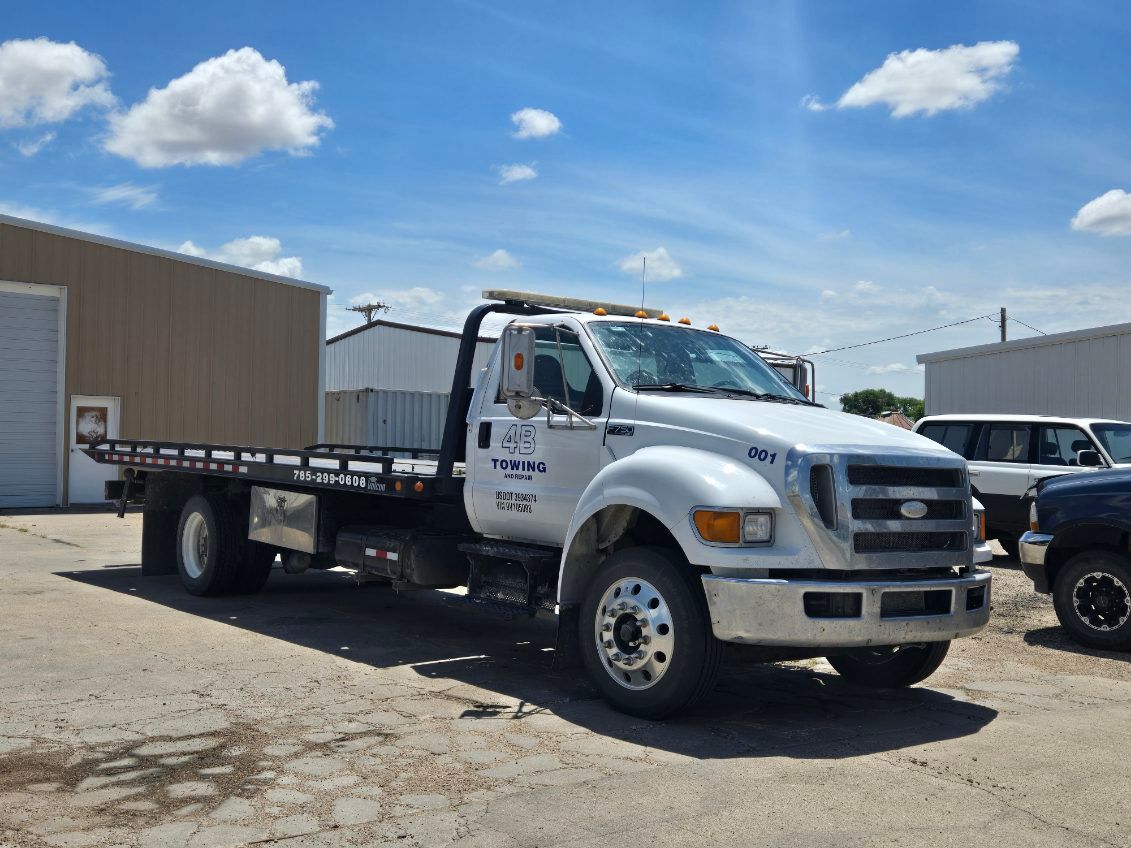 A white Ford flatbed tow truck sits parked on a concrete lot outside commercial buildings under a sunny blue sky. The vehicle features "4B Towing" branding on its side door.
