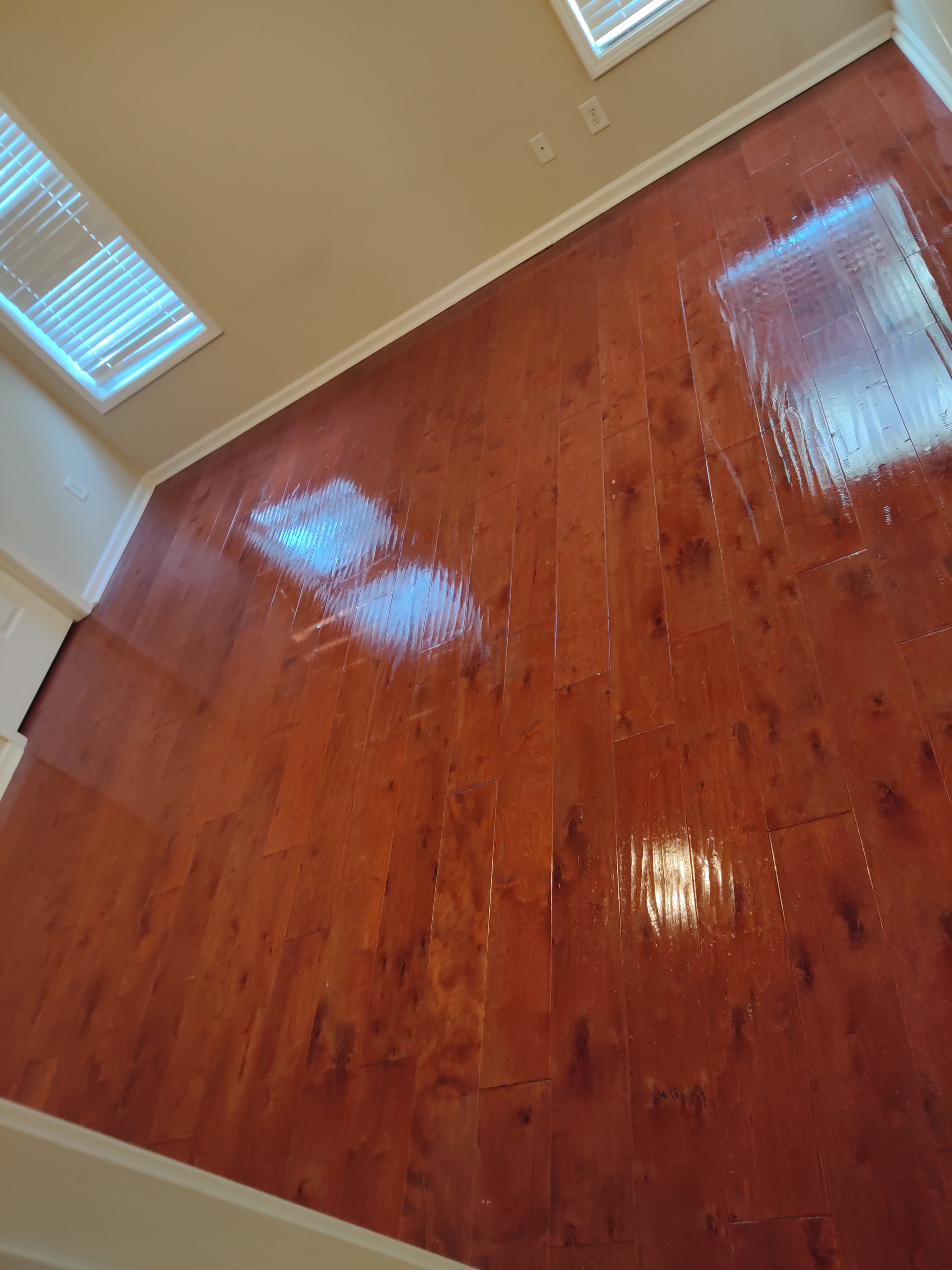 A high-angle view of a polished, reddish-brown hardwood floor reflecting bright light from ceiling windows and possibly other light sources. The floor is made of planks and appears to be recently cleaned or waxed. The walls are a neutral, light beige color, and there are white baseboards. Two electrical outlets are visible on the wall.