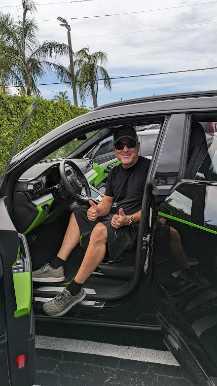 A man in a black t-shirt, black shorts, and a black baseball cap is sitting in the driver's seat of a black Lamborghini Urus. The car has neon green accents on the interior. The man is smiling and giving a double thumbs up. The car is parked outdoors, with palm trees and a leafy green hedge visible in the background.
