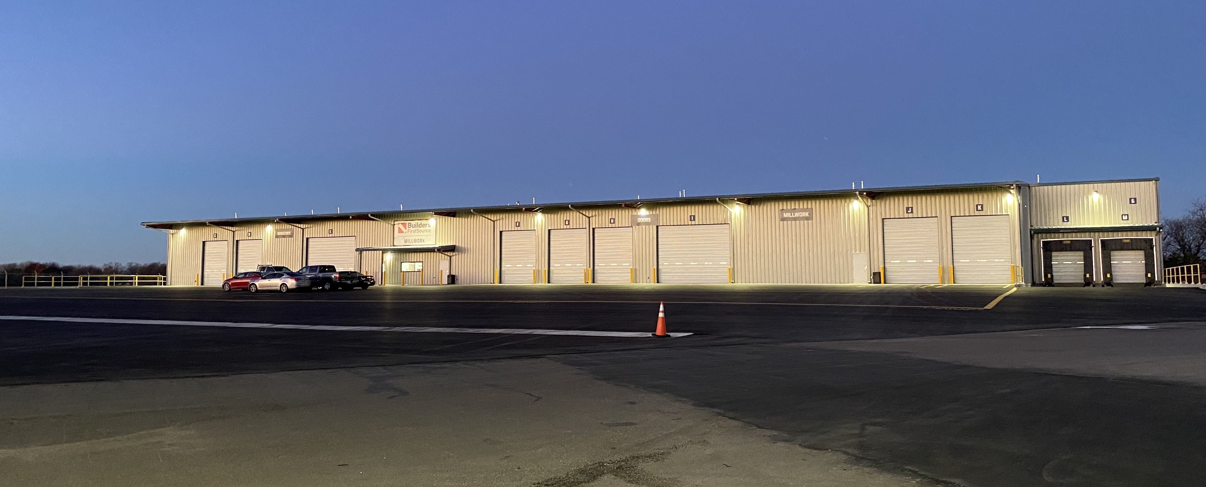 Exterior view of a large Builders FirstSource millwork facility at dusk, featuring multiple illuminated loading bays labeled for doors and millwork. The metal-clad building stands against a clear evening sky, with a few parked vehicles and a traffic cone in the foreground, highlighting the spacious and well-lit lot.