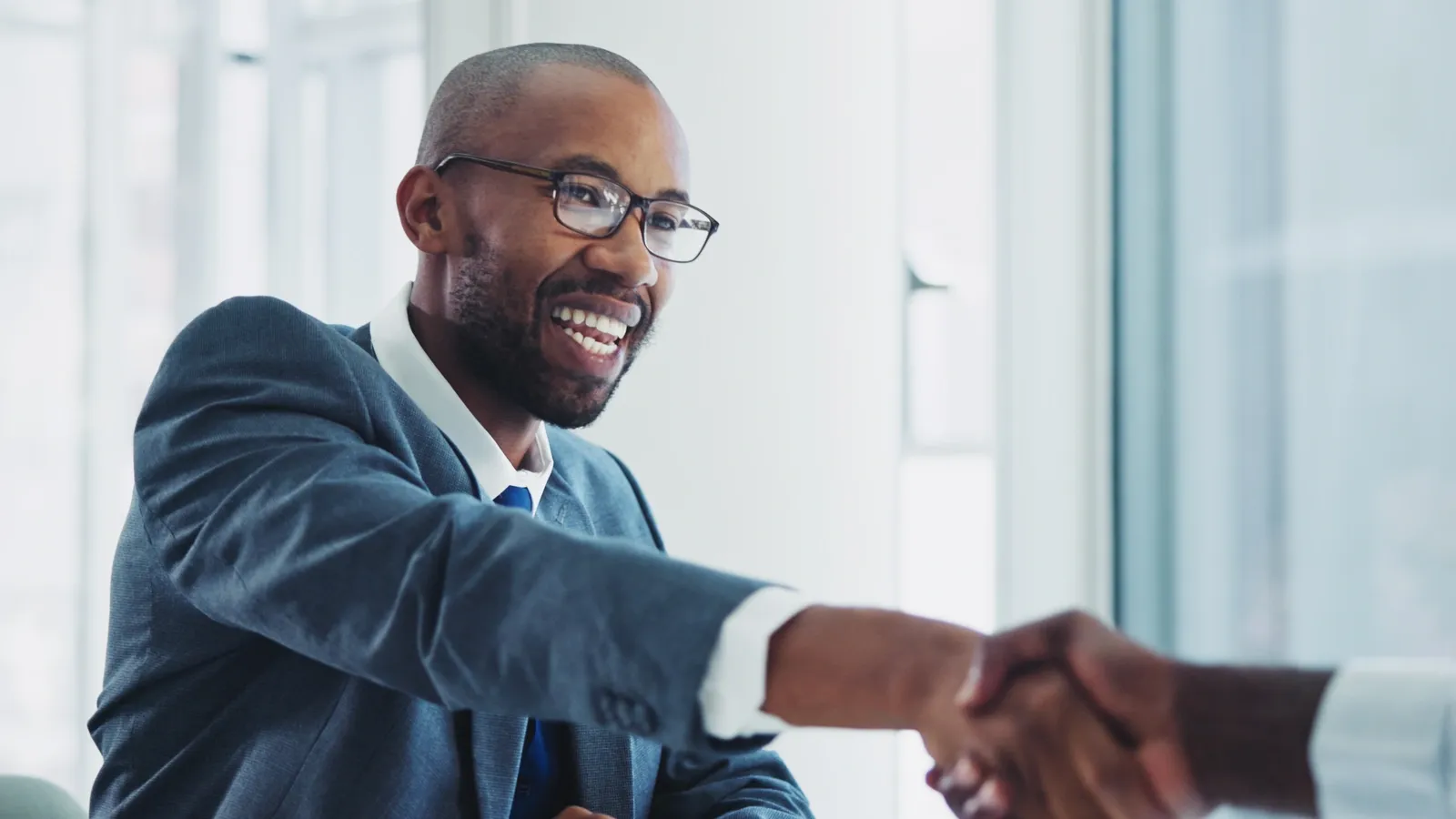 A man wearing a suit and glasses smiles broadly while shaking hands with another person. The background is blurred and appears to be an office setting with natural light.