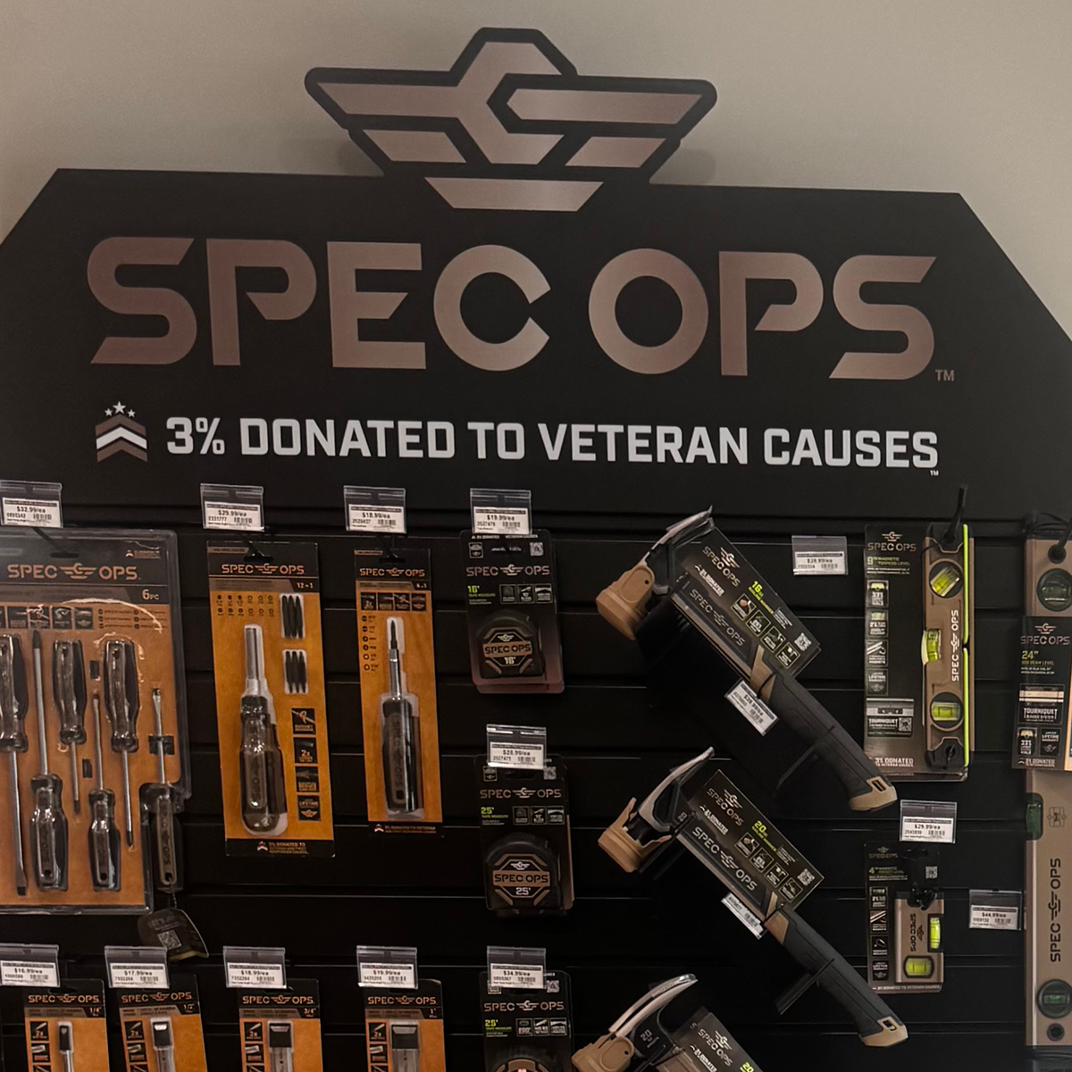 A black slatwall display in a Builders FirstSource store features Spec Ops hand tools and accessories in black-and-orange packaging. Several tape measures, utility knives, screwdrivers, levels, and other tools hang below a large Spec Ops logo. Text beneath the logo reads “3% donated to veteran causes.”
