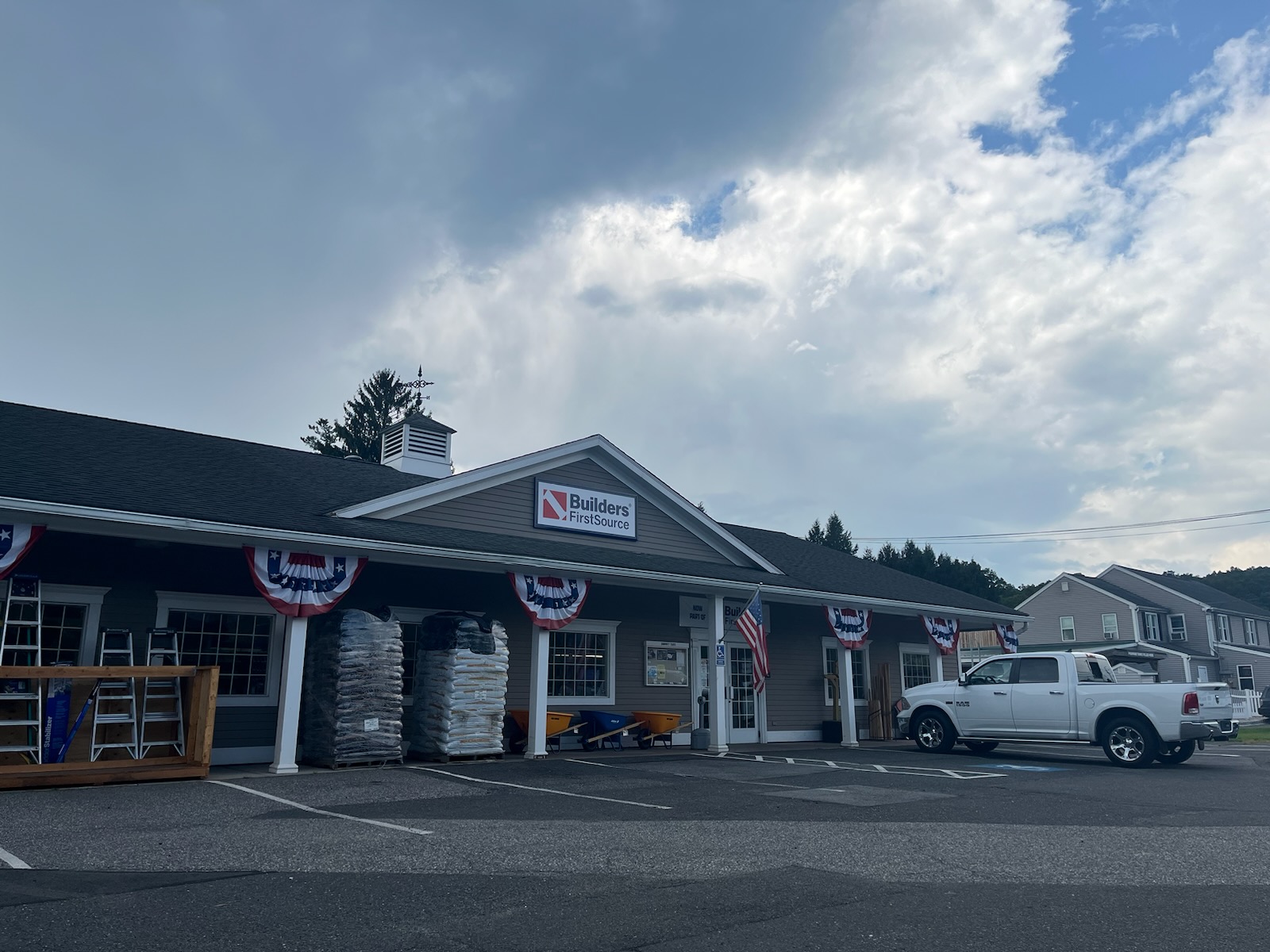 Exterior view of a Builders FirstSource store in Oxford, CT, with a white pickup truck parked outside. The storefront is decorated with patriotic bunting under a cloudy sky. Building materials, including stacked bags and ladders, are displayed outside the store. The store's sign is prominently displayed on the roof.