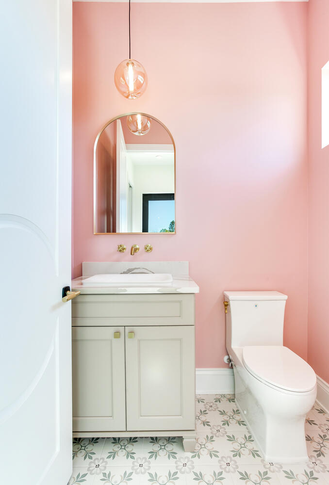Charming powder bathroom with soft pink walls, a light vanity, arched mirror, brass lighting, and stylish patterned floor tile.