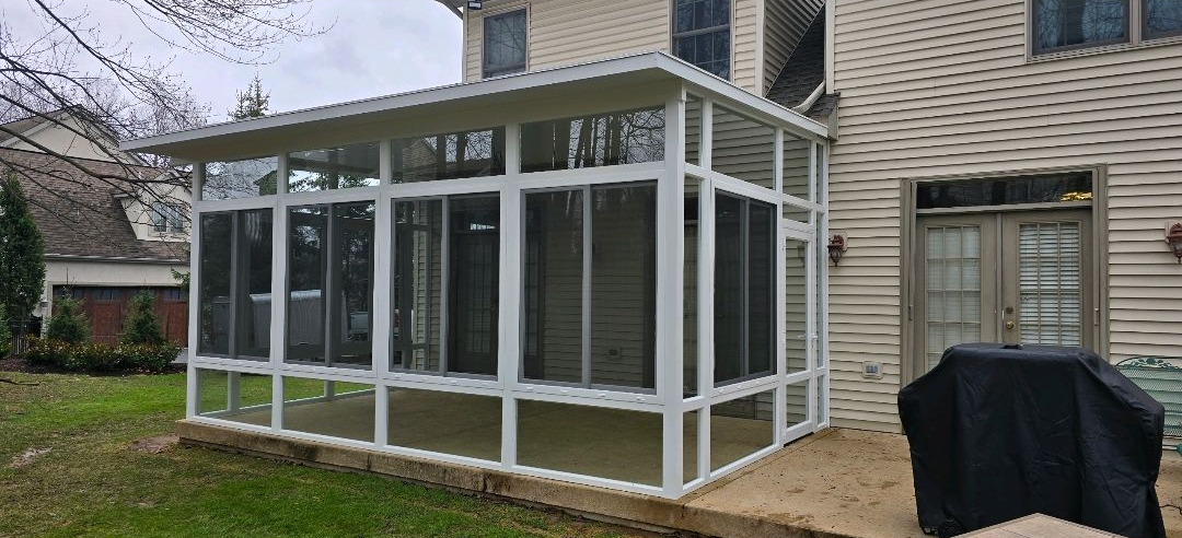 Modern screened-in patio extension attached to a house, featuring white framing, large mesh panels, and a flat roof. The structure provides a spacious, enclosed outdoor area, ideal for relaxing while enjoying fresh air without exposure to insects.
