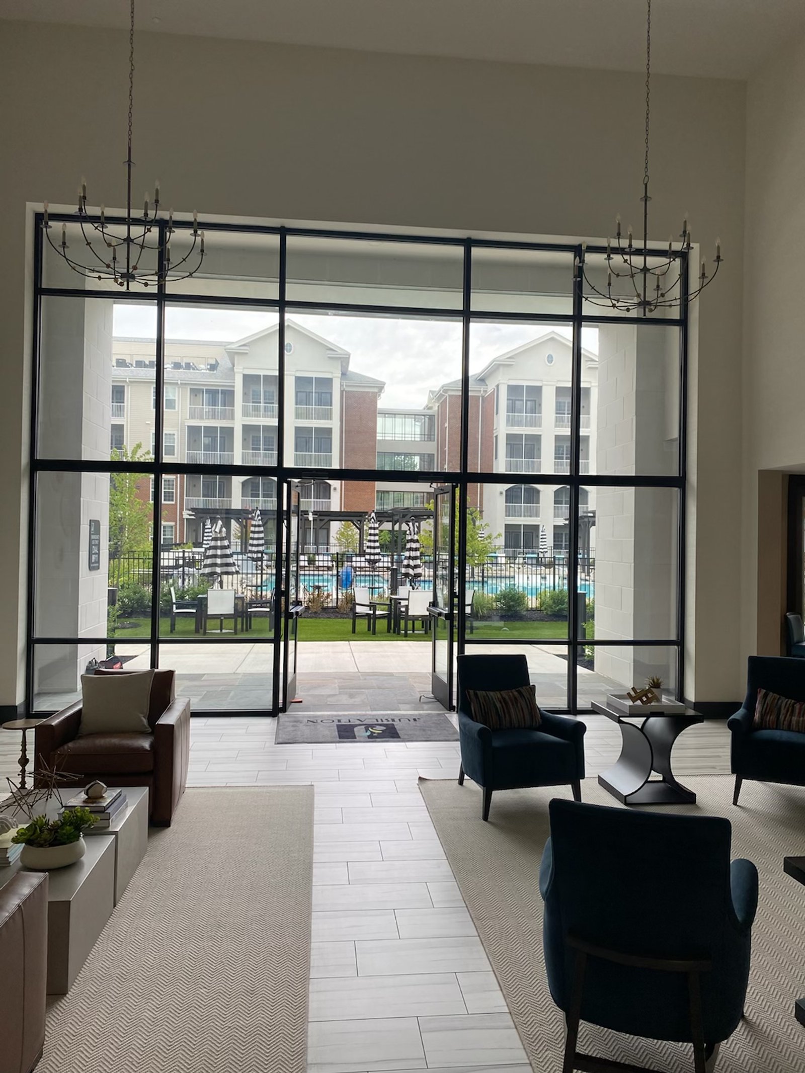 A high-ceilinged lobby area with large windows overlooking an apartment complex pool and outdoor seating area. The lobby features modern furniture including leather armchairs, plush blue armchairs, and contemporary coffee tables. Two ornate chandeliers hang from the ceiling. The view through the window shows a swimming pool, lounge chairs, umbrellas, and the exterior of the apartment buildings.