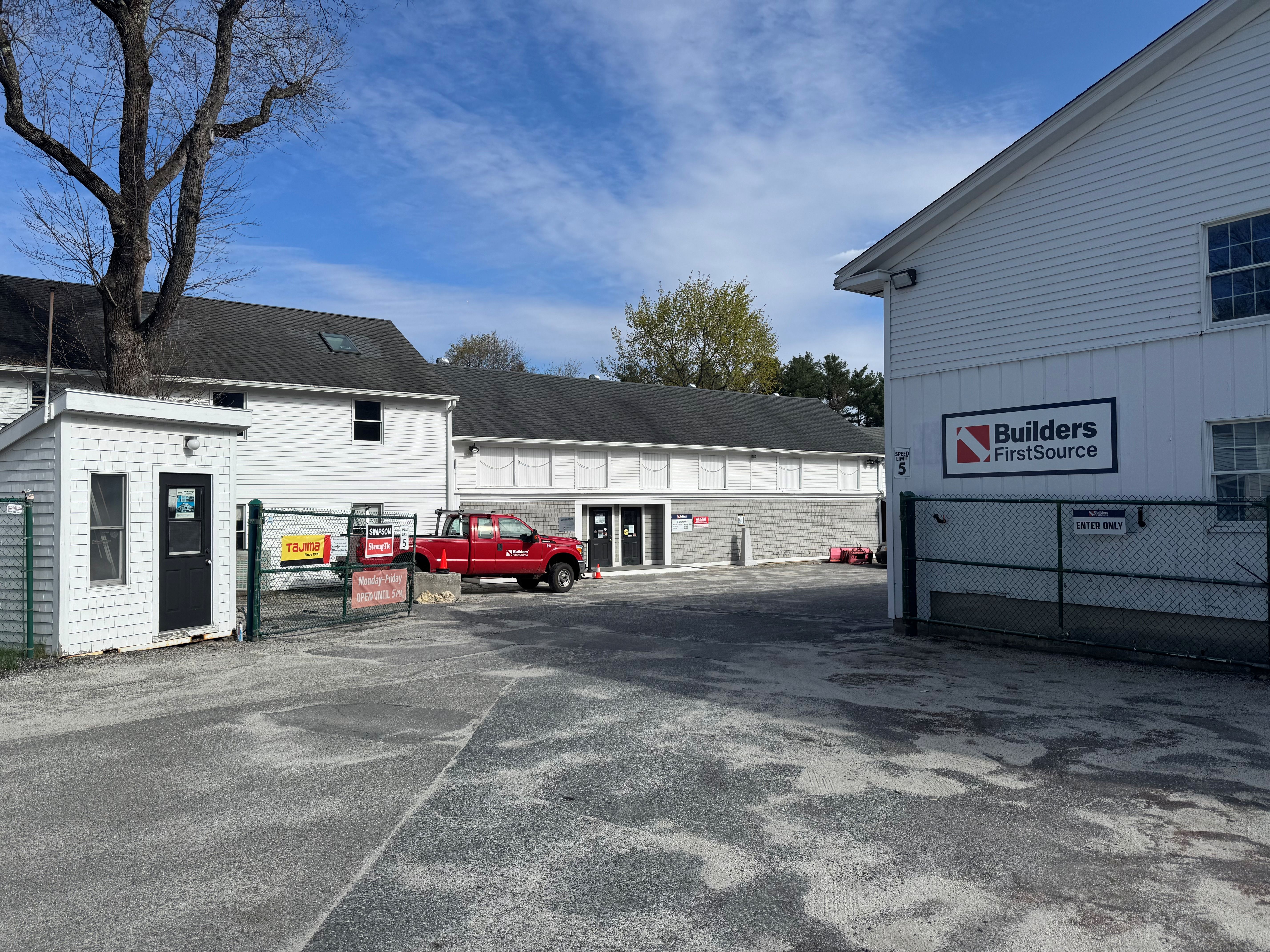 Exterior view of a Builders FirstSource facility with white wood-sided buildings, a red pickup truck parked near service doors, and a paved open yard under a blue sky.