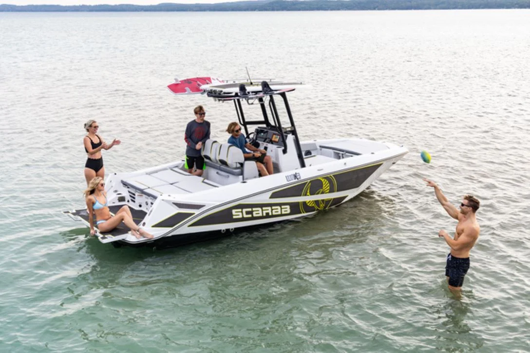 A group of four people enjoy a day on a Scarab boat in calm water. Two women are sitting on the back of the boat, one in a black bikini and the other in a blue bikini. Two men are on the boat, one steering and the other standing. Another man is in the water to the right of the boat, throwing a colorful ball.