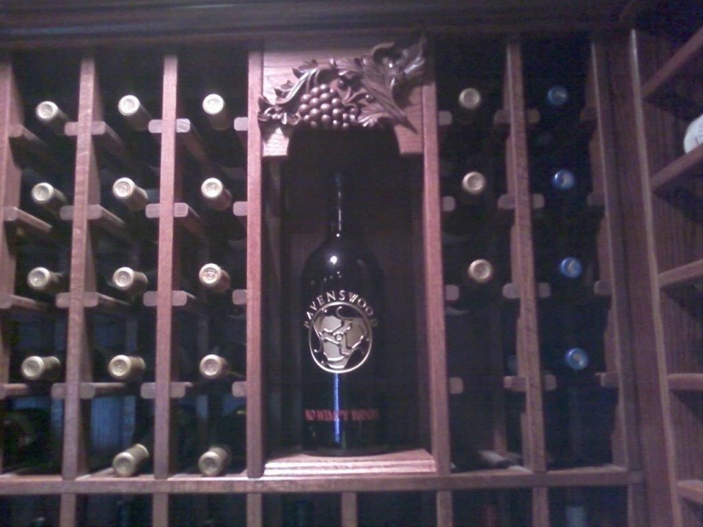A wooden wine rack displaying multiple bottles arranged horizontally, with a decorative central section highlighting a large wine bottle and carved grape design, showcasing elegant storage and presentation.