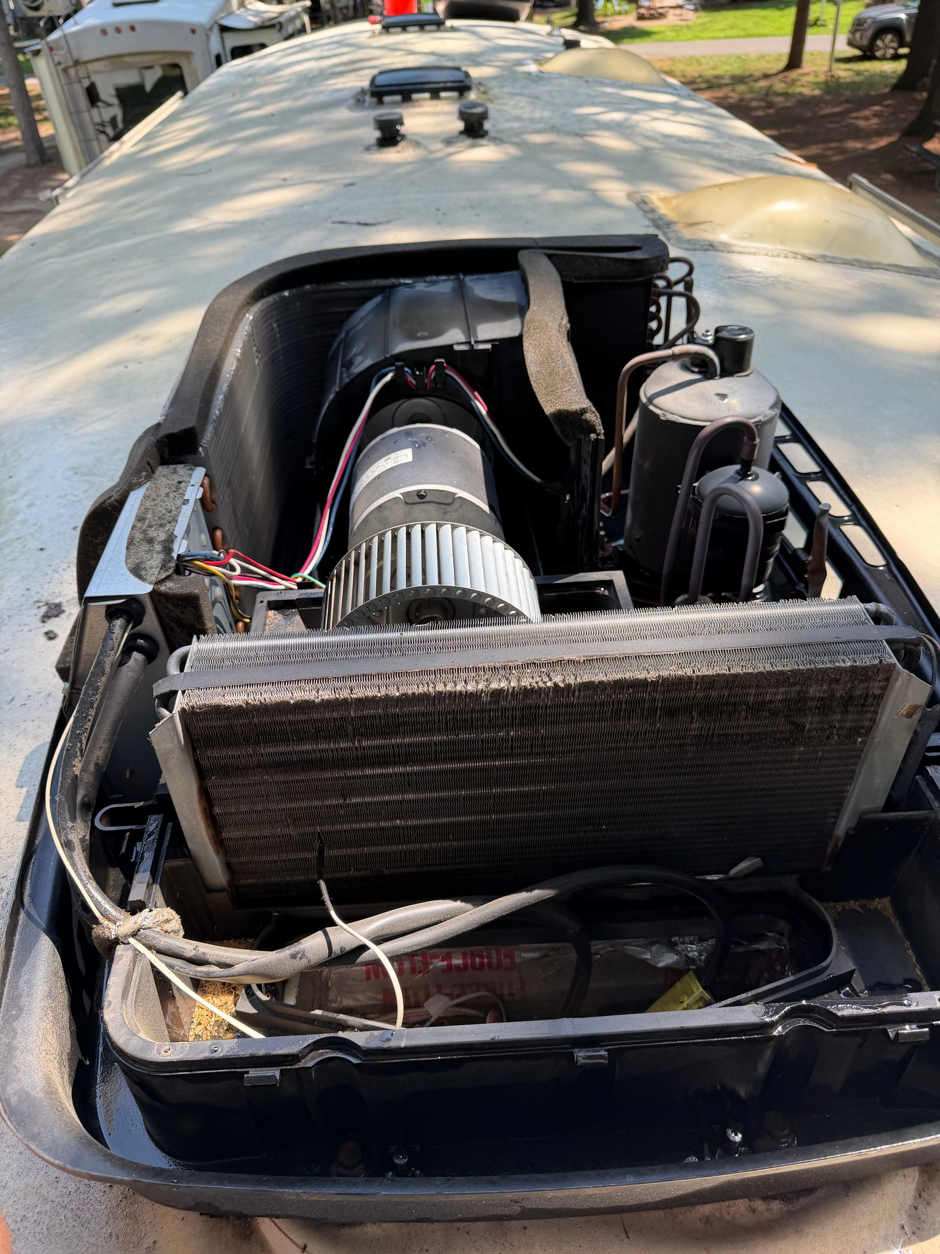 Open rooftop RV air-conditioning compartment reveals condenser coils, blower motor, wiring, refrigerant lines, compressor components, and surrounding housing, showing the internal mechanical system during inspection or repair work outdoors nearby.