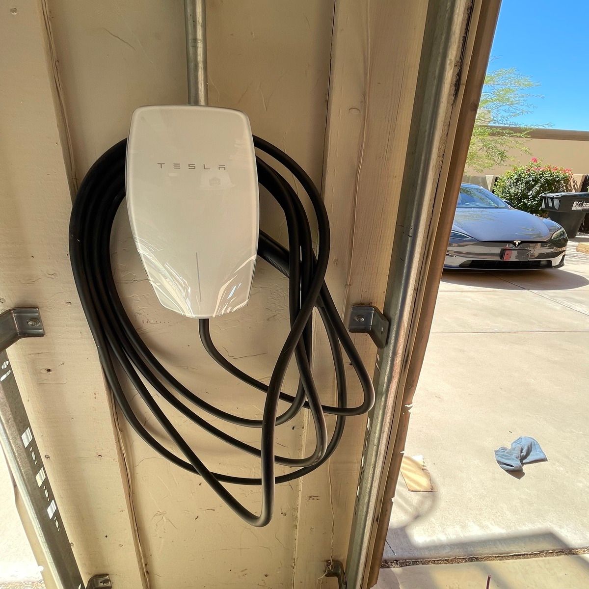 A Tesla wall connector is mounted inside a residential garage with its thick black charging cable neatly looped around the unit, while a Tesla vehicle remains parked outside in sunlight.