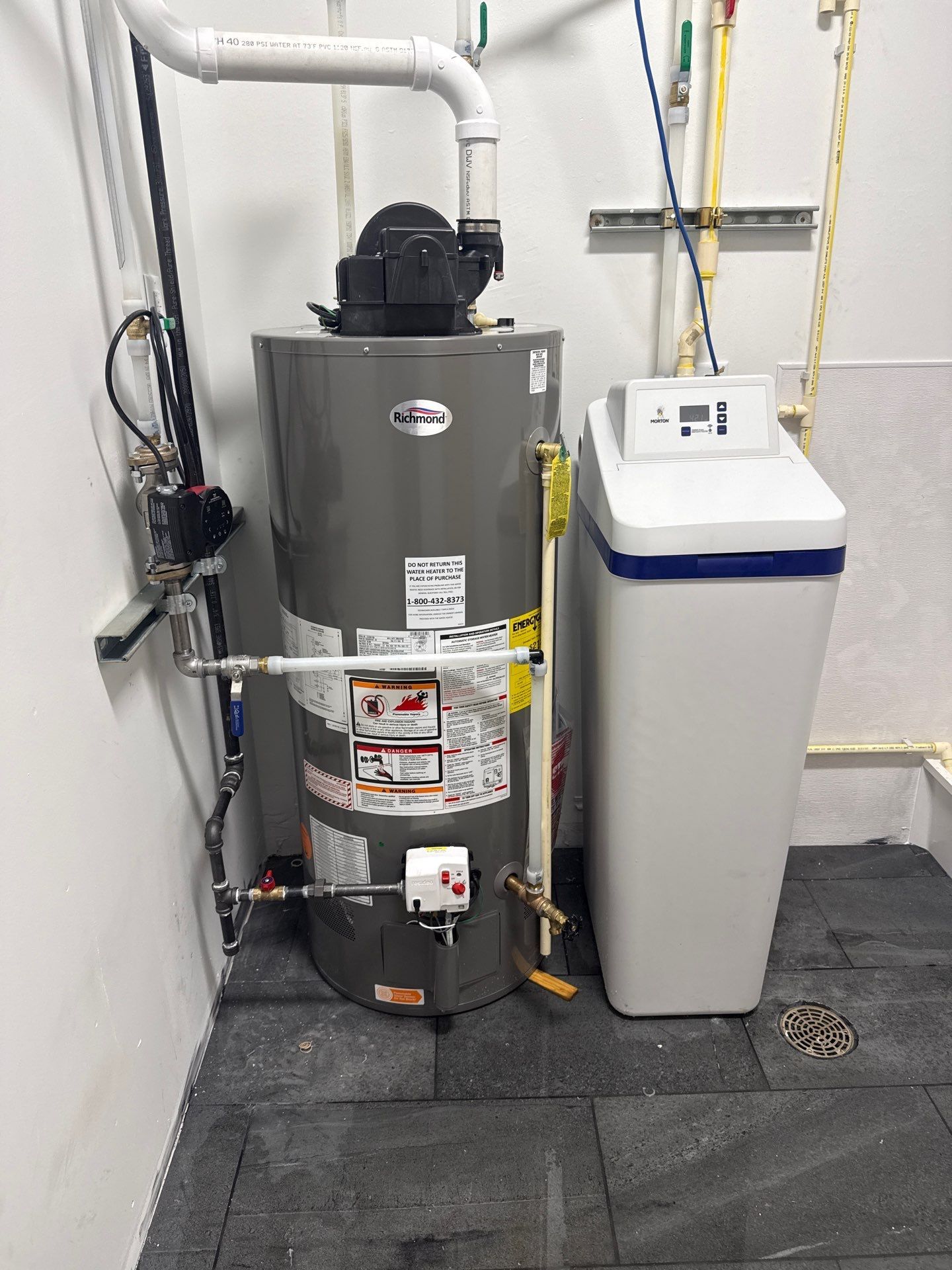 Gray water heater and white water softener installed in a utility room, connected with PVC, copper, and flexible piping, valves, controls, warning labels, and nearby floor drain for household plumbing.