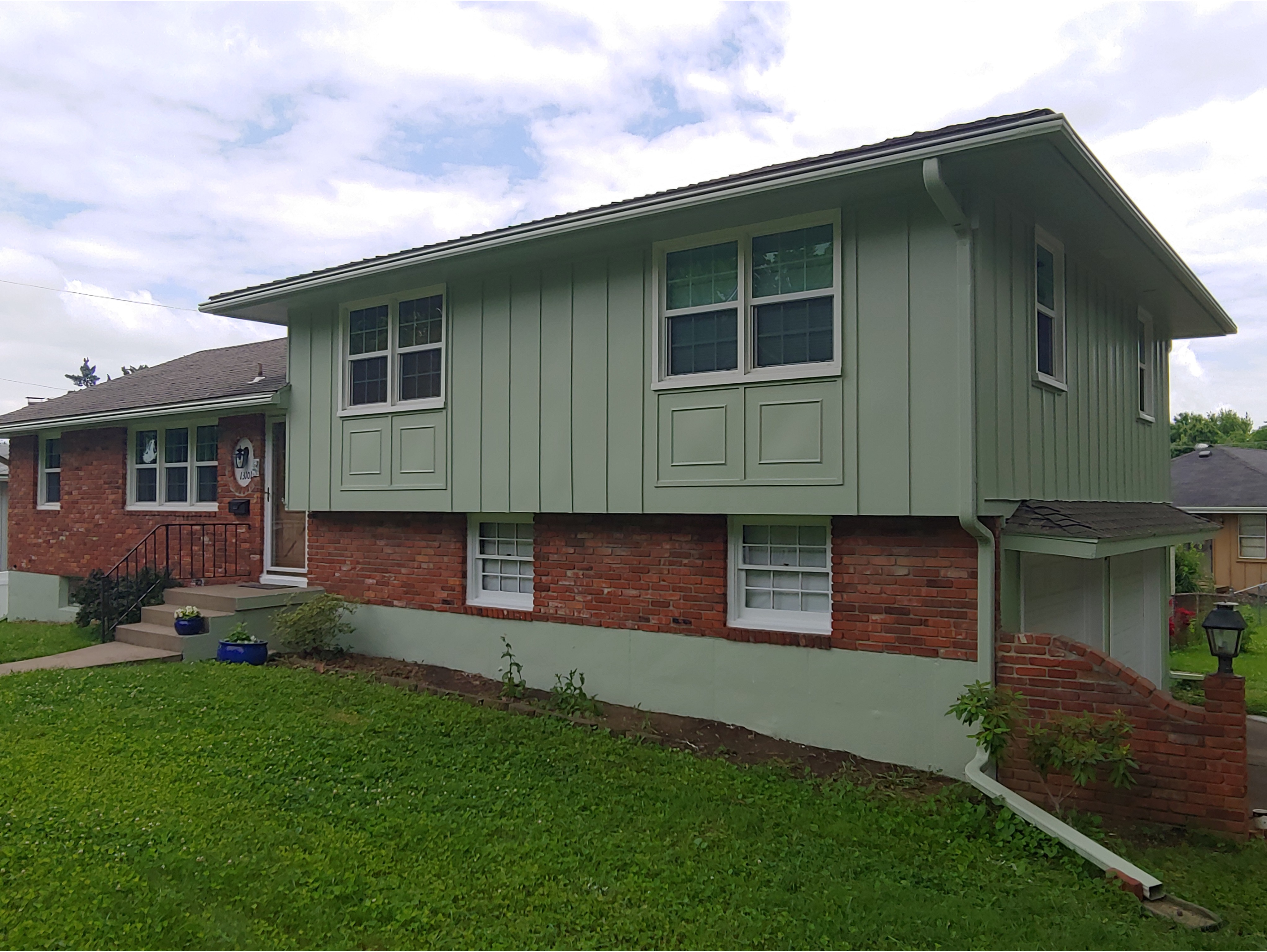 A split-level home featuring a fresh exterior paint job with light green siding and red brick accents. The house has white-trimmed windows and a well-maintained lawn, complemented by a neatly landscaped entryway with potted plants. Ideal for showcasing professional exterior painting services and home renovation transformations.