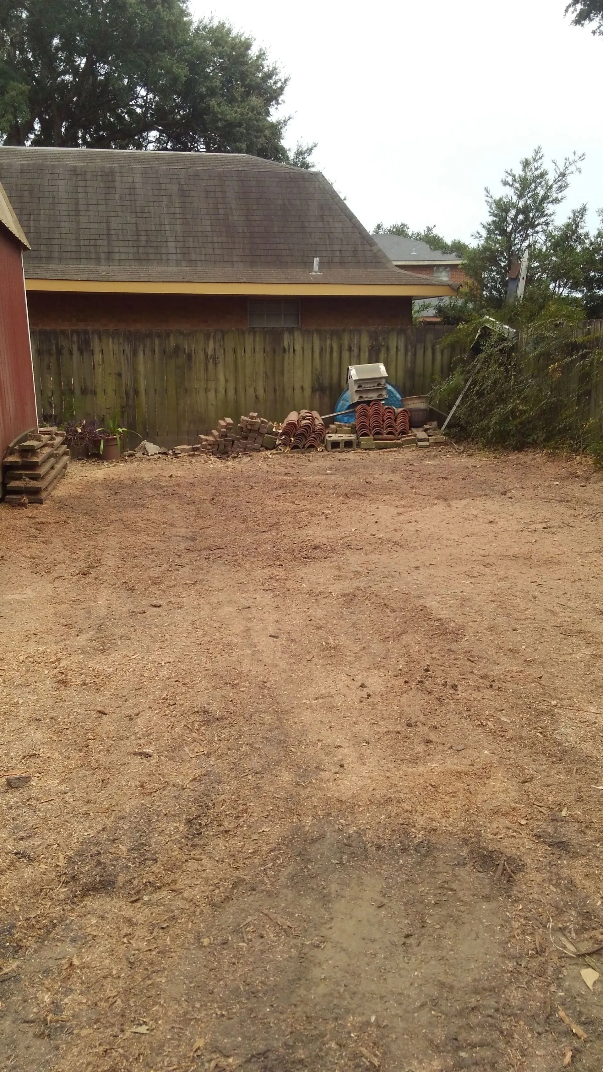 A cleared dirt yard with scattered sawdust, stacked bricks, tiles, and a grill in front of a wooden fence.