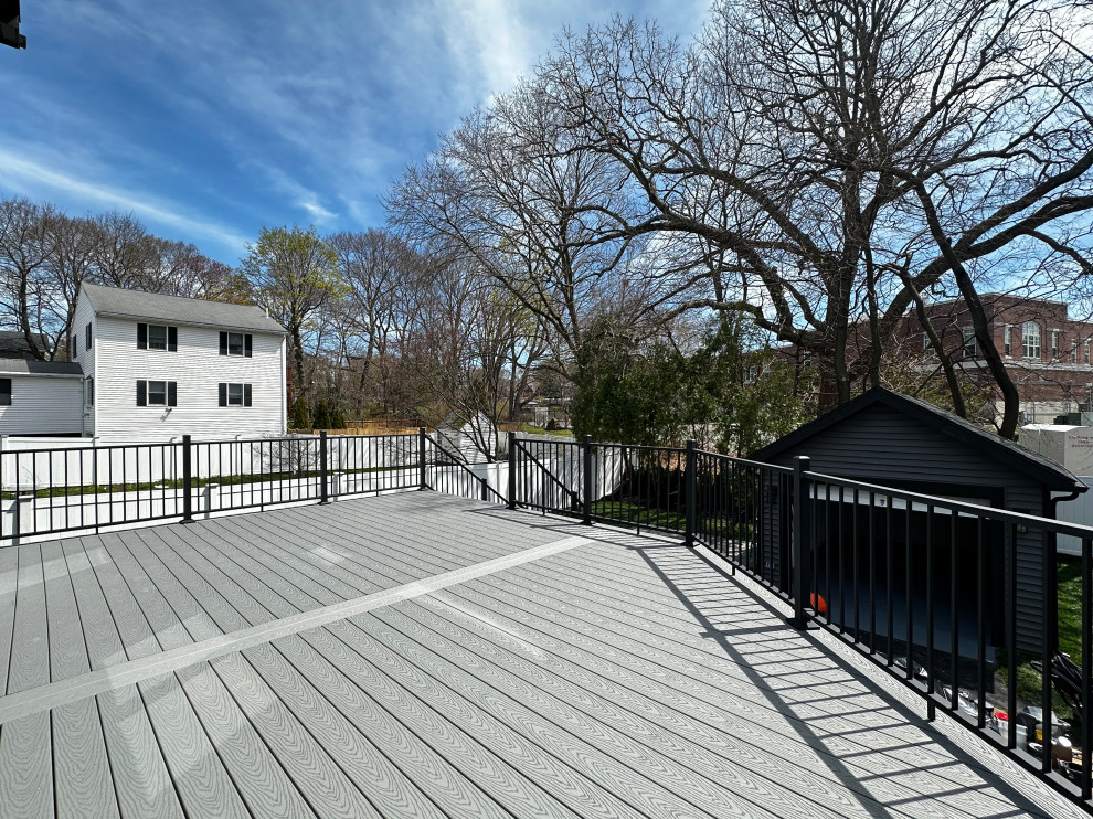 A large, modern grey composite deck with black railings overlooks a suburban neighborhood with houses, trees, and a small dark shed. The sky is blue with scattered white clouds and the sun is casting shadows from the railing onto the deck.