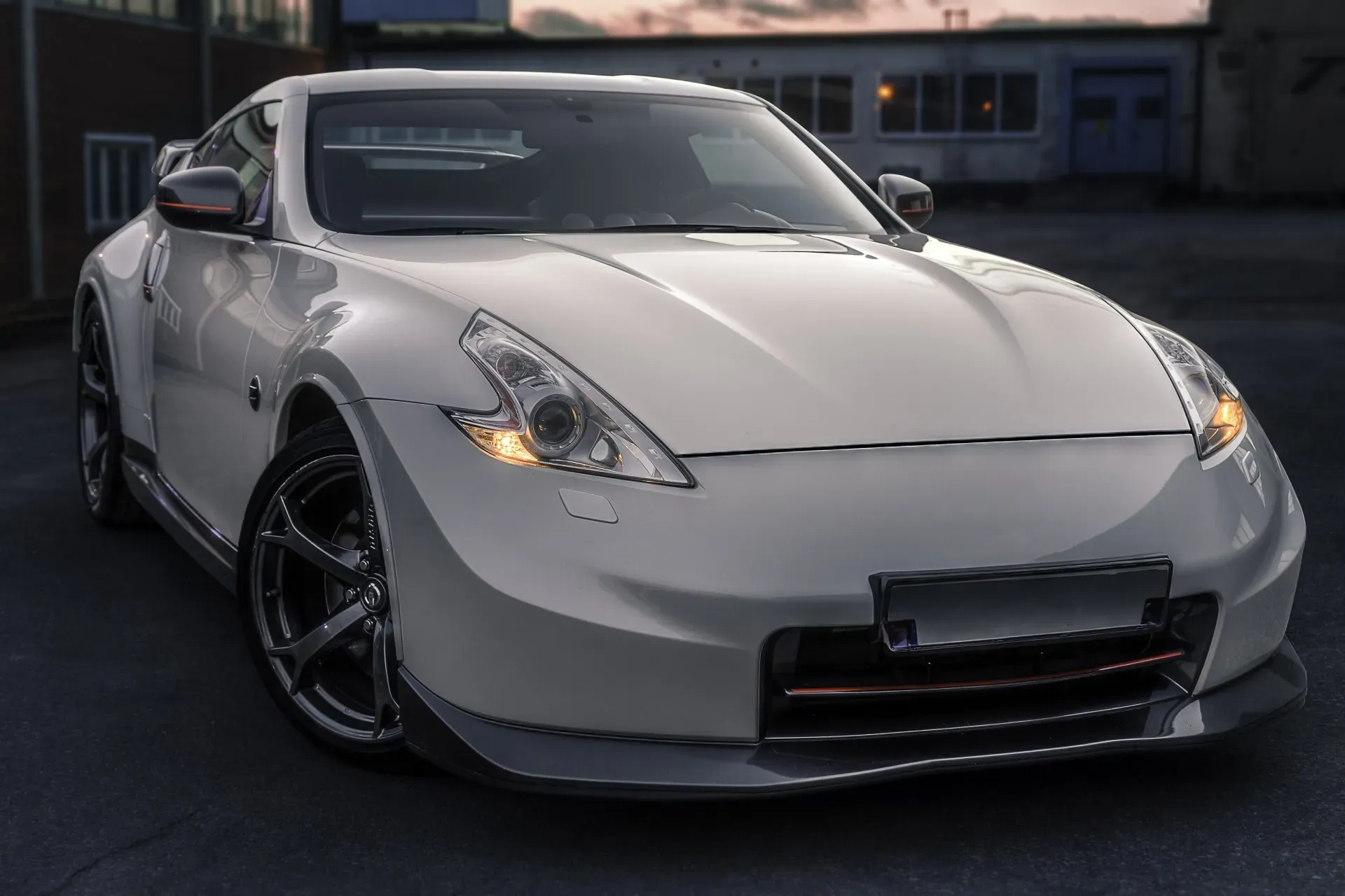 A white Nissan 370Z sports car is parked on a dark asphalt surface in front of a building with windows. The car has its headlights on and features a sleek, aerodynamic design with dark rims and a lower body kit. The background shows a muted sky with hints of sunset and the facade of an industrial or garage-like building.