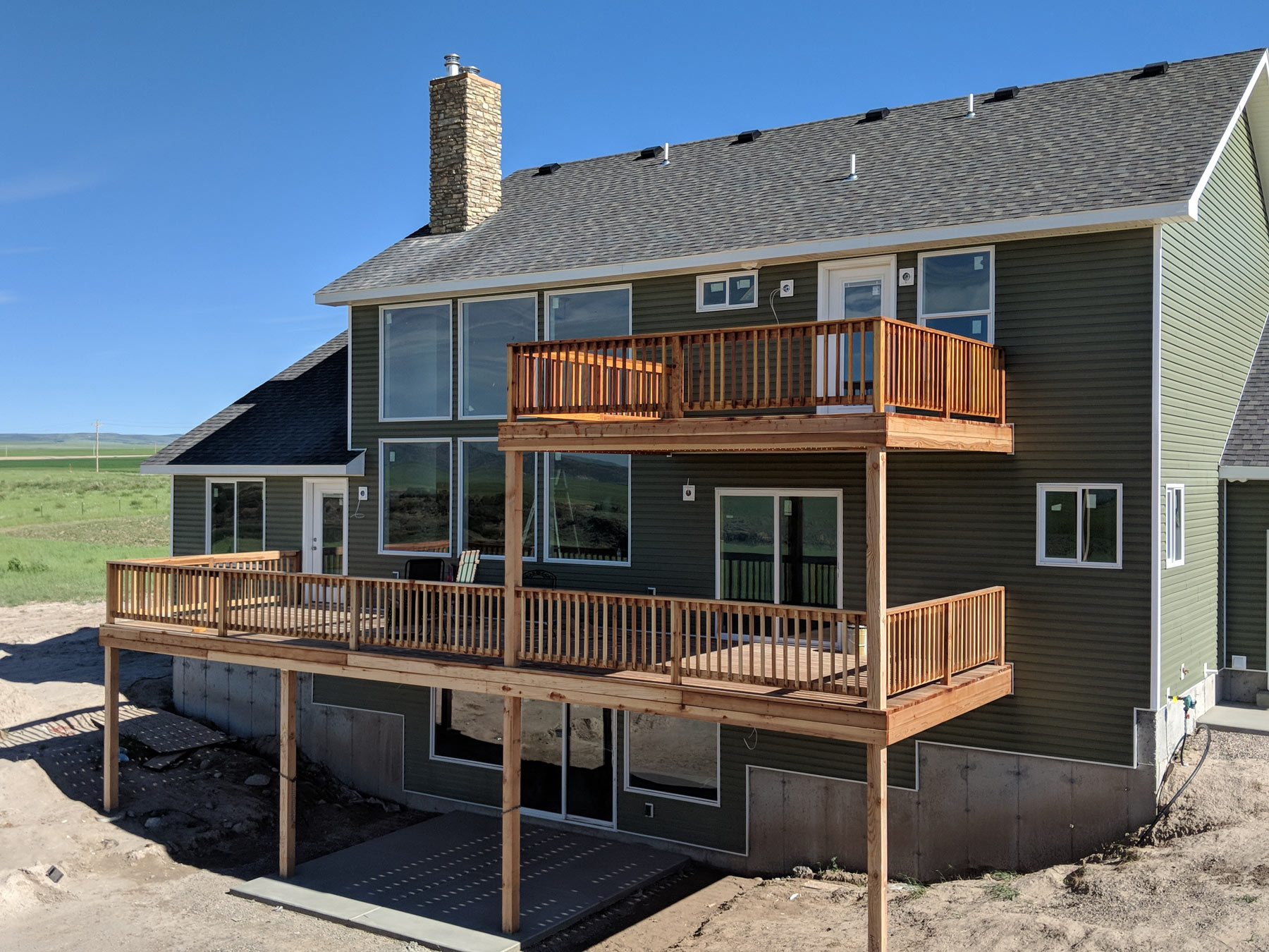 Enhance your new construction with a stunning multi-level natural wood deck by DeckPro LLC. This custom design features an upper-level balcony accessible from the master suite and a expansive lower-level deck perfect for entertaining, all with safe and stylish wood railings. The warm wood tones create a perfect contrast with the deep olive green siding, integrating beautifully with the stone veneer chimney and surrounding open landscape.