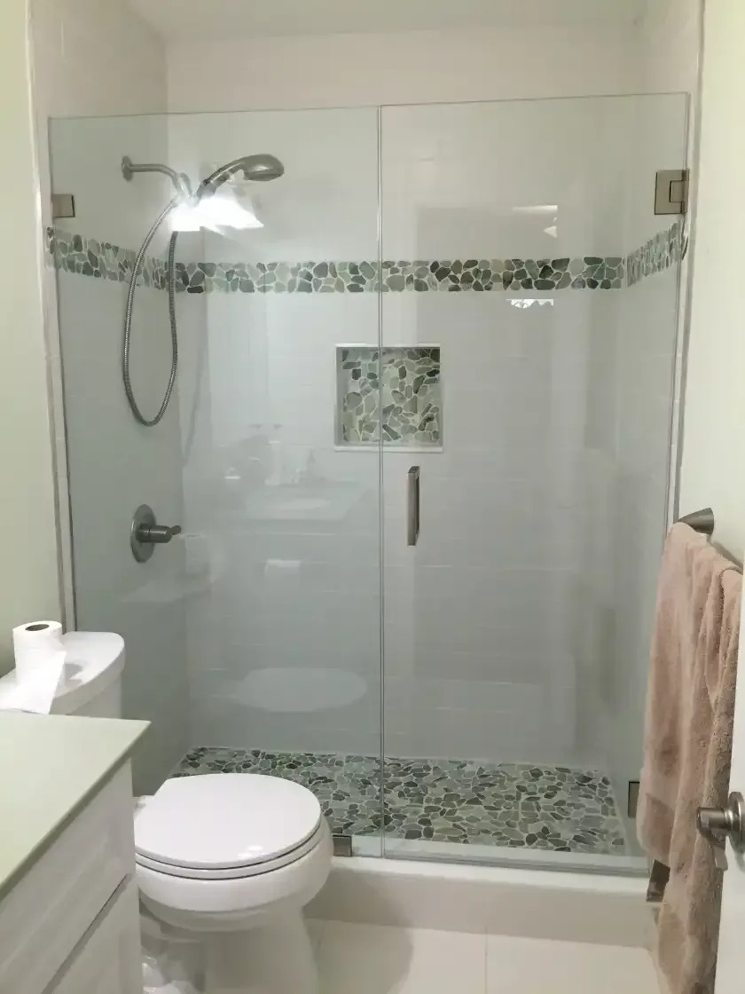A modern bathroom features a glass-enclosed shower with horizontally striped gray and white tile. The shower head and faucet are silver, and blue and pink sponges hang inside.