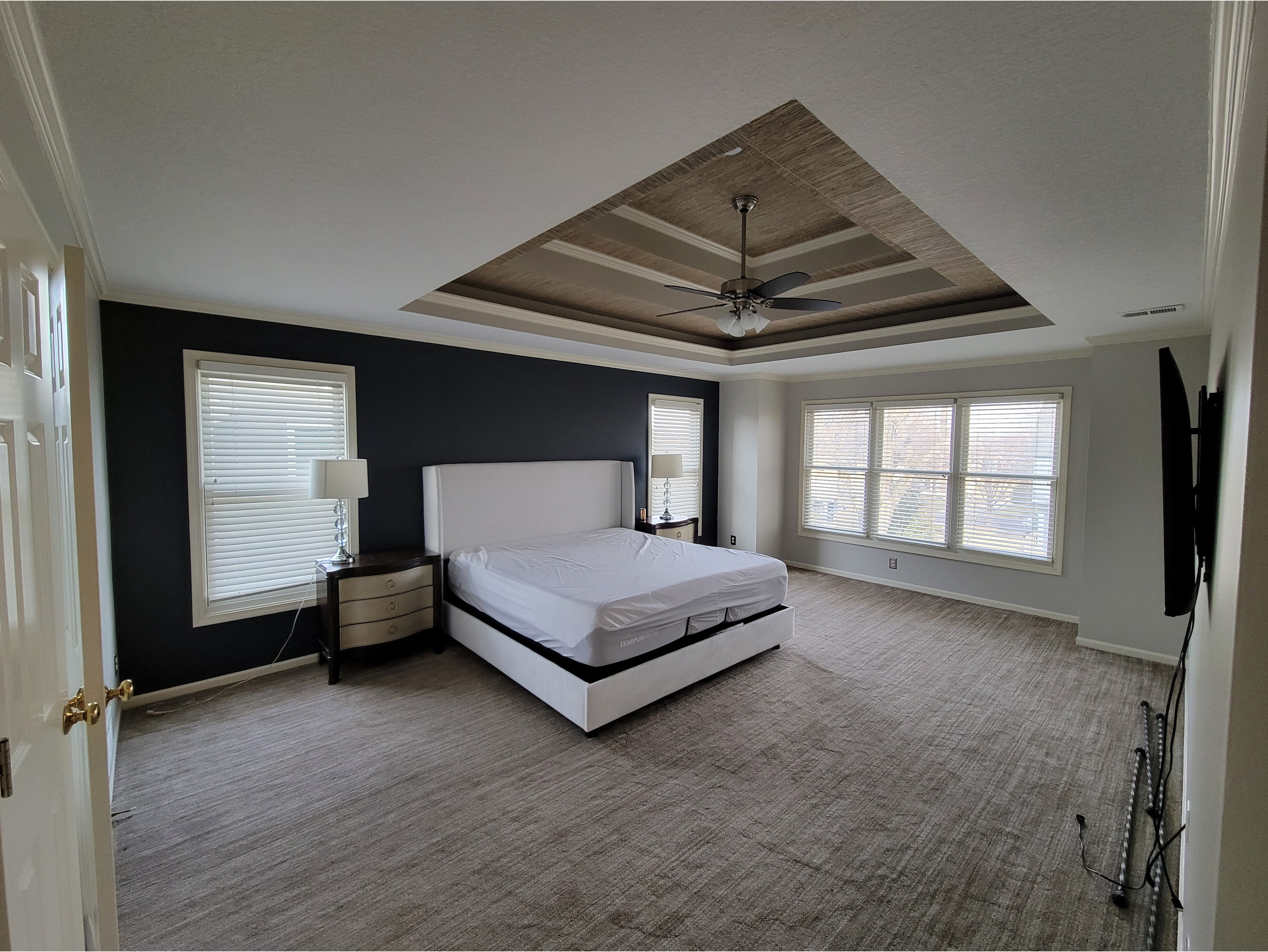 A modern master bedroom featuring a fresh interior paint job with a dark accent wall and light neutral tones for contrast. The room includes a tray ceiling with custom detailing and a ceiling fan, adding style and dimension. Large windows with white trim provide ample natural light. Perfect for showcasing interior painting services, accent wall designs, and ceiling enhancements.