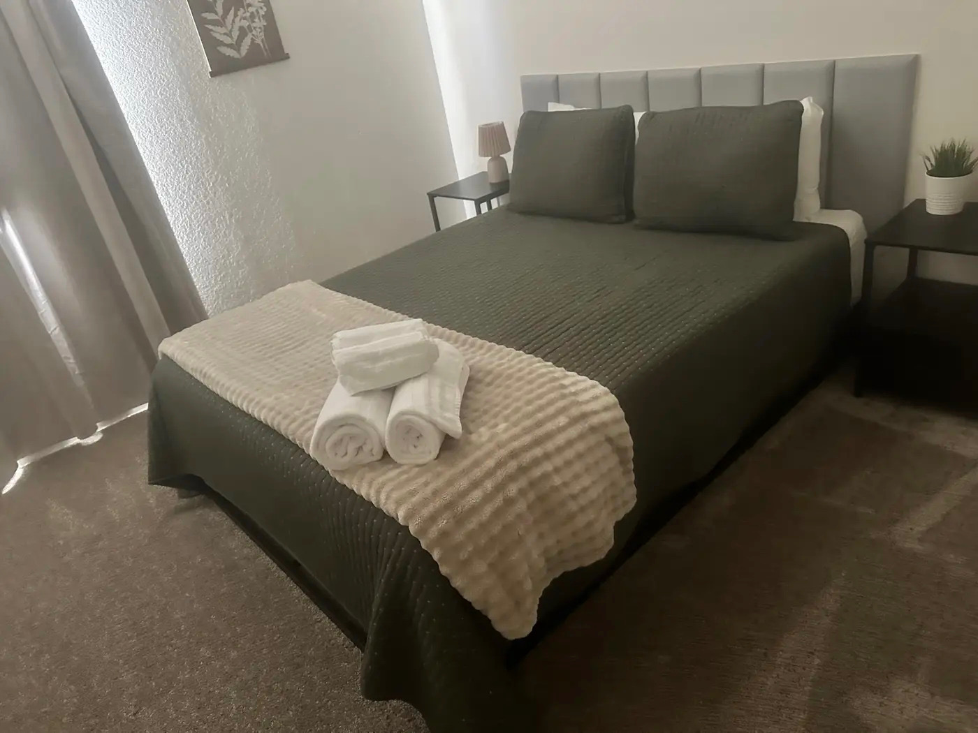 A neatly made bed in a bedroom, with a grey quilted comforter, a grey upholstered headboard, and two dark green pillows. White towels are folded and placed on top of a beige textured throw blanket on the bed. A bedside table with a lamp and a small potted plant is visible on the right side of the bed. The room has grey curtains on the left and a framed piece of art on the wall.