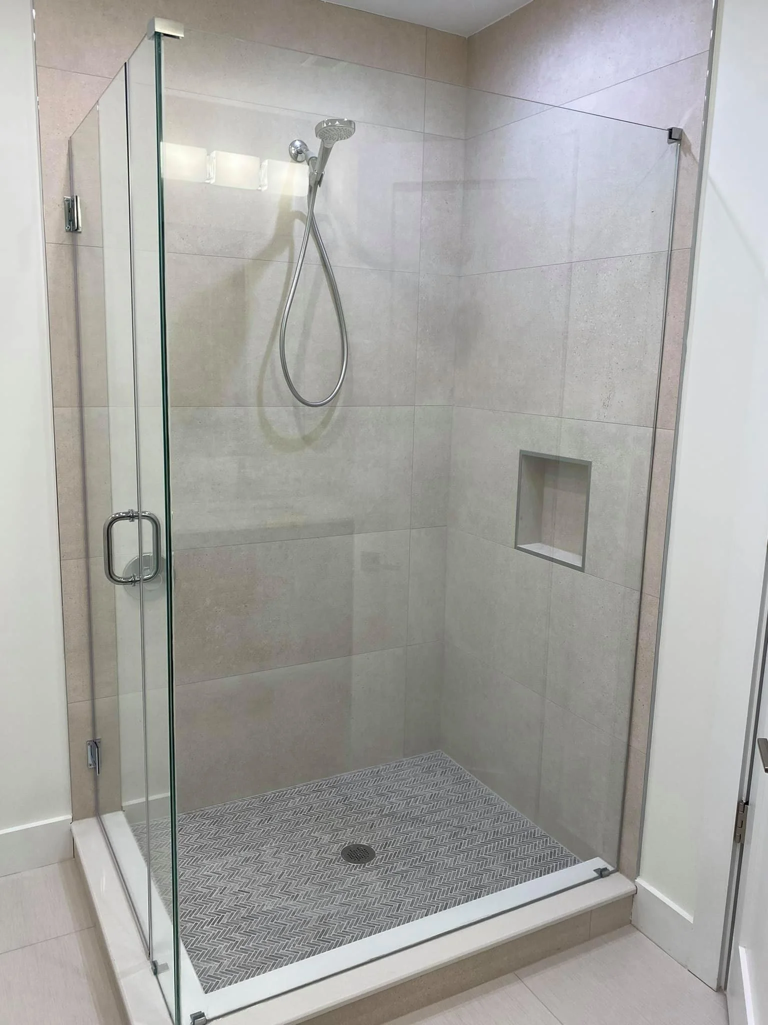 A modern, frameless glass shower enclosure with a chrome handle and showerhead is shown. The walls are tiled with light gray square tiles, and the floor has a herringbone pattern mosaic. A recessed niche is visible on the back wall, and the enclosure is situated within a clean, well-lit bathroom.