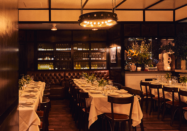 An elegant, dimly lit private dining space features long tables set for a group. Dark wood walls and a large wine display case create a sophisticated, upscale ambiance.