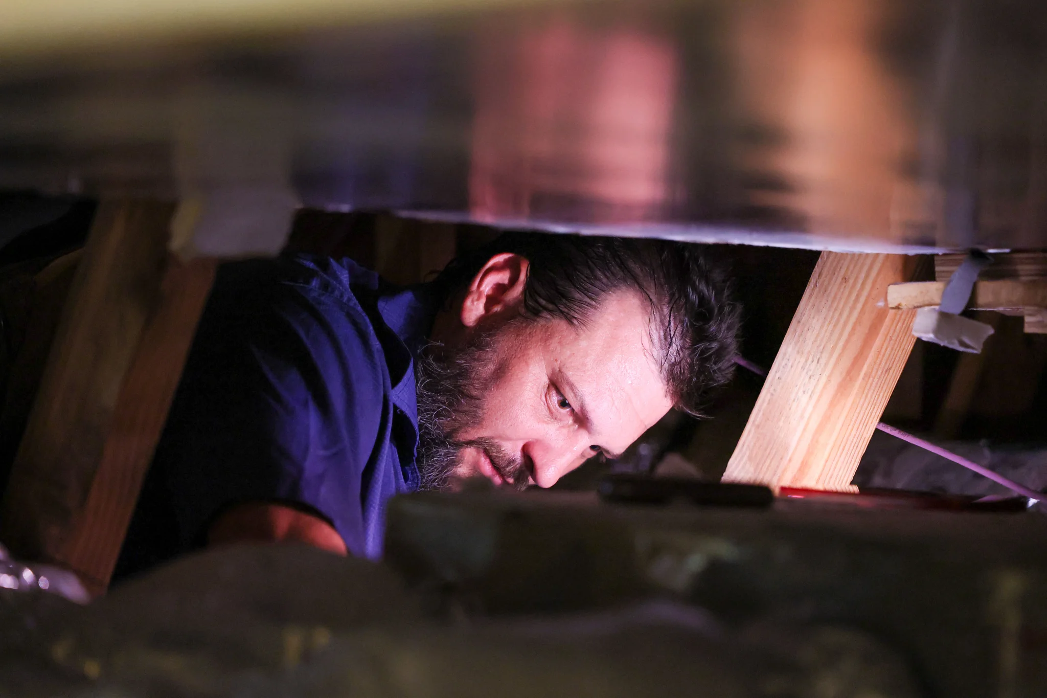 A man with a beard wearing a blue shirt works in a cramped attic space, surrounded by wooden beams a 