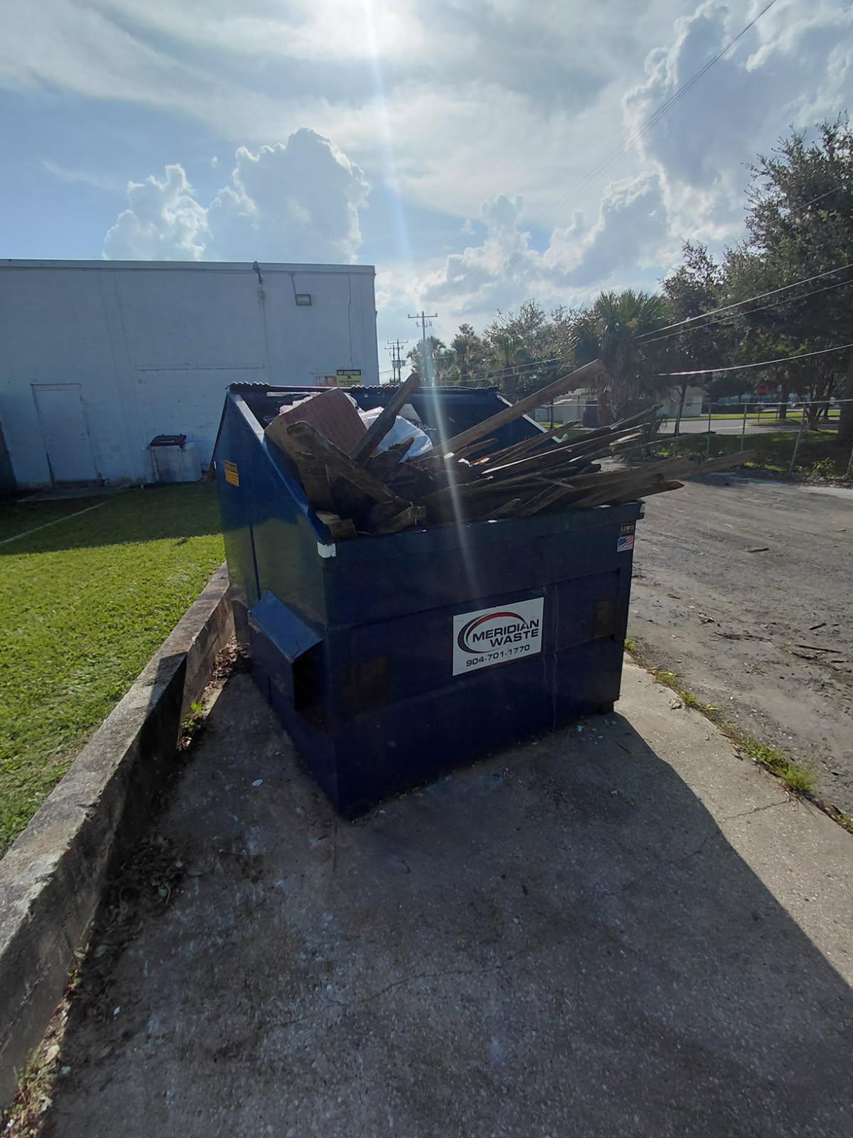 A blue Meridian Waste dumpster overflowing with discarded wooden planks and construction debris. 