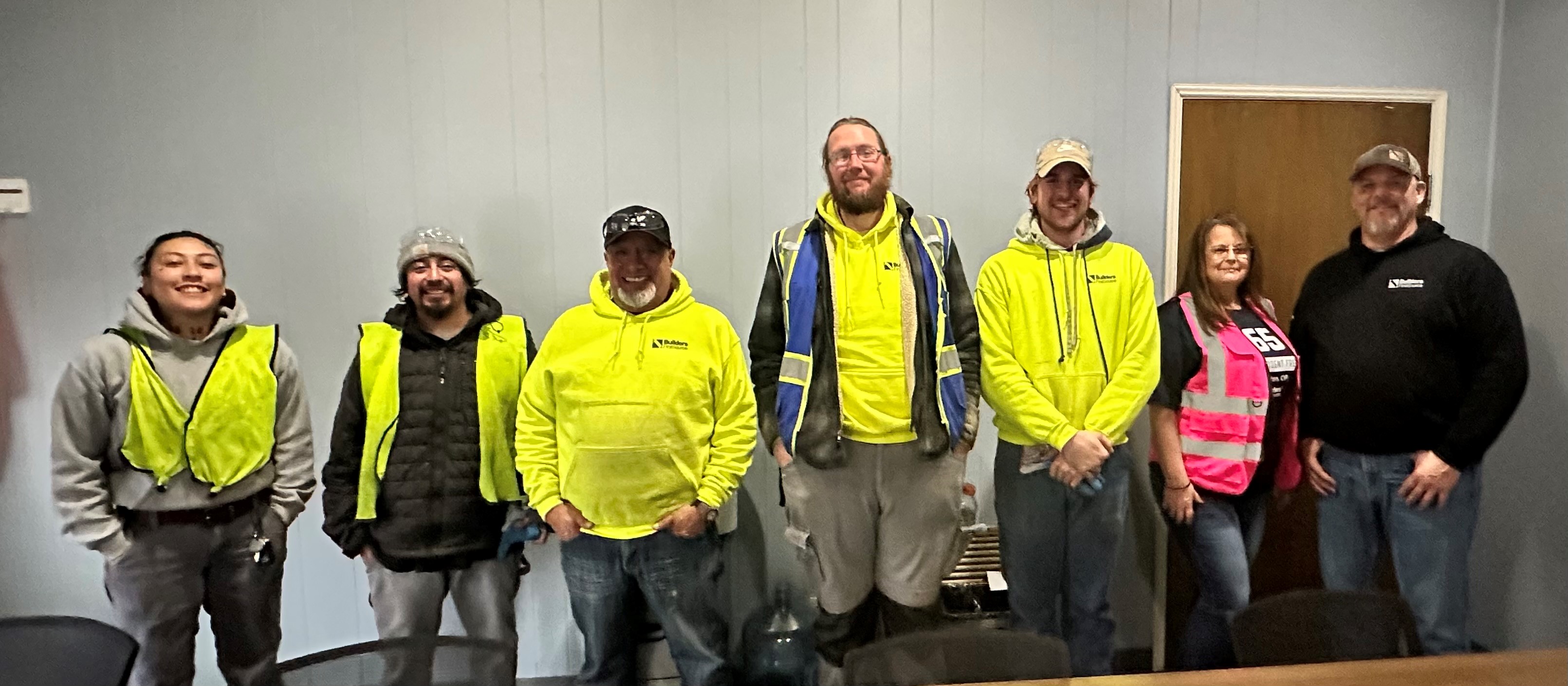 A group of seven employees are standing together, smiling for a group photo in an indoor office setting. Most of the individuals are wearing high-visibility safety vests and jackets. The group includes both men and women, with one woman wearing a bright pink vest that says "Safety Committee." The others are in various shades of yellow and blue safety gear, with one person wearing a beanie and another in a cap. They appear relaxed and friendly, standing against a plain wall with a door visible behind them on the right side of the image.