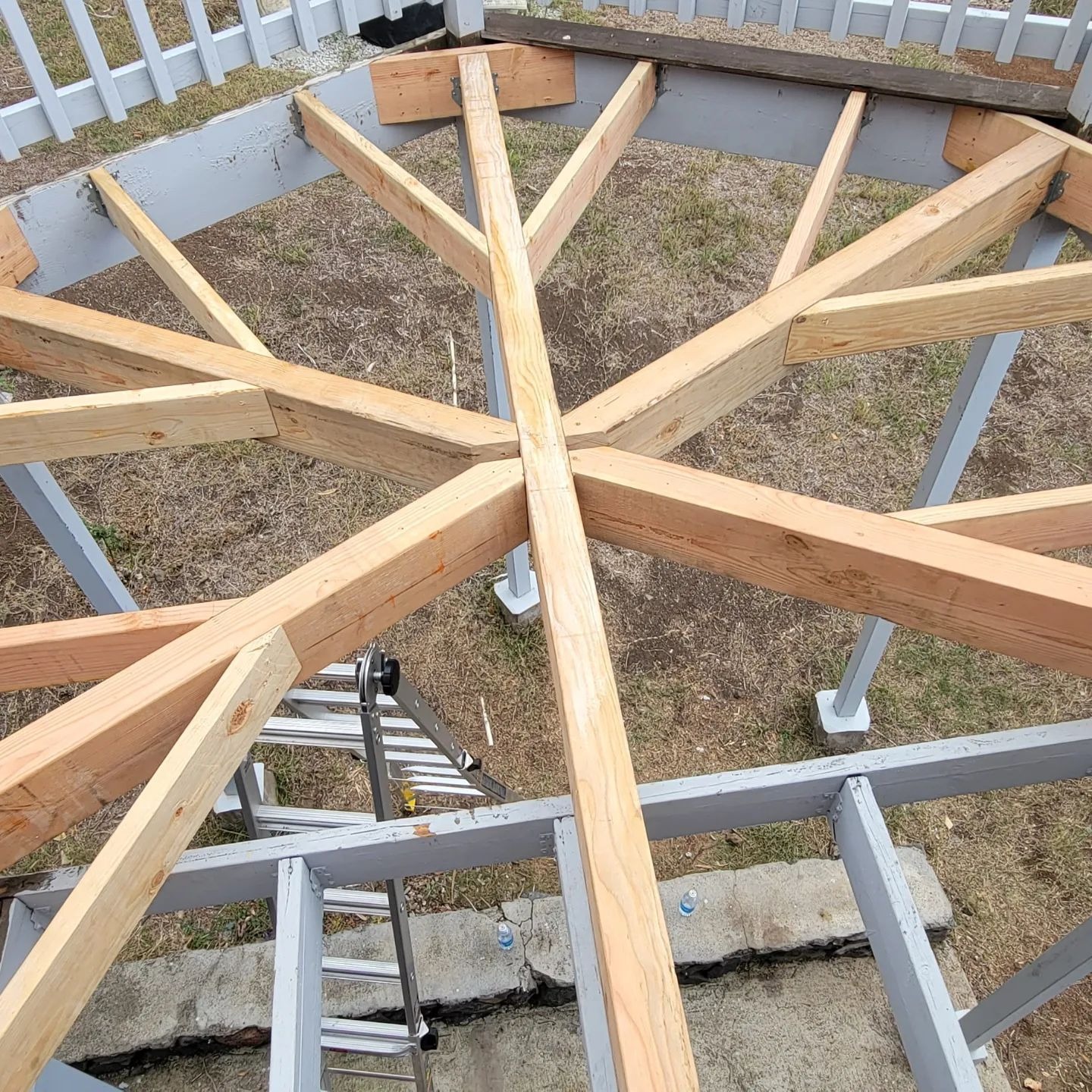 An overhead view of the wooden framing for a hexagonal or circular deck under construction. Multiple wooden joists radiate from a central point, supported by painted gray beams and posts on a grassy lawn.