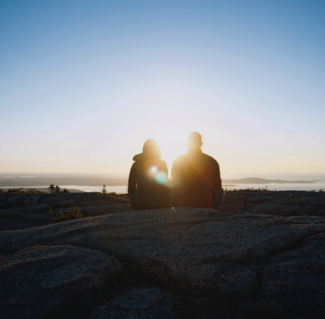 Two people sitting together on a hill, silhouetted by the setting sun.