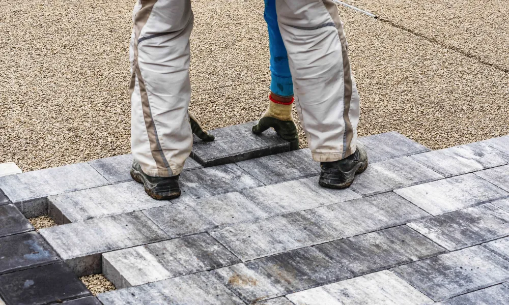 A close-up, low-angle view of two people laying down gray interlocking pavers on a gravel base. The pavers have a marbled pattern of light and dark gray. The individuals are wearing work pants, gloves, and work boots. The background is out of focus, showing more gravel and a hint of a finished paver section.
