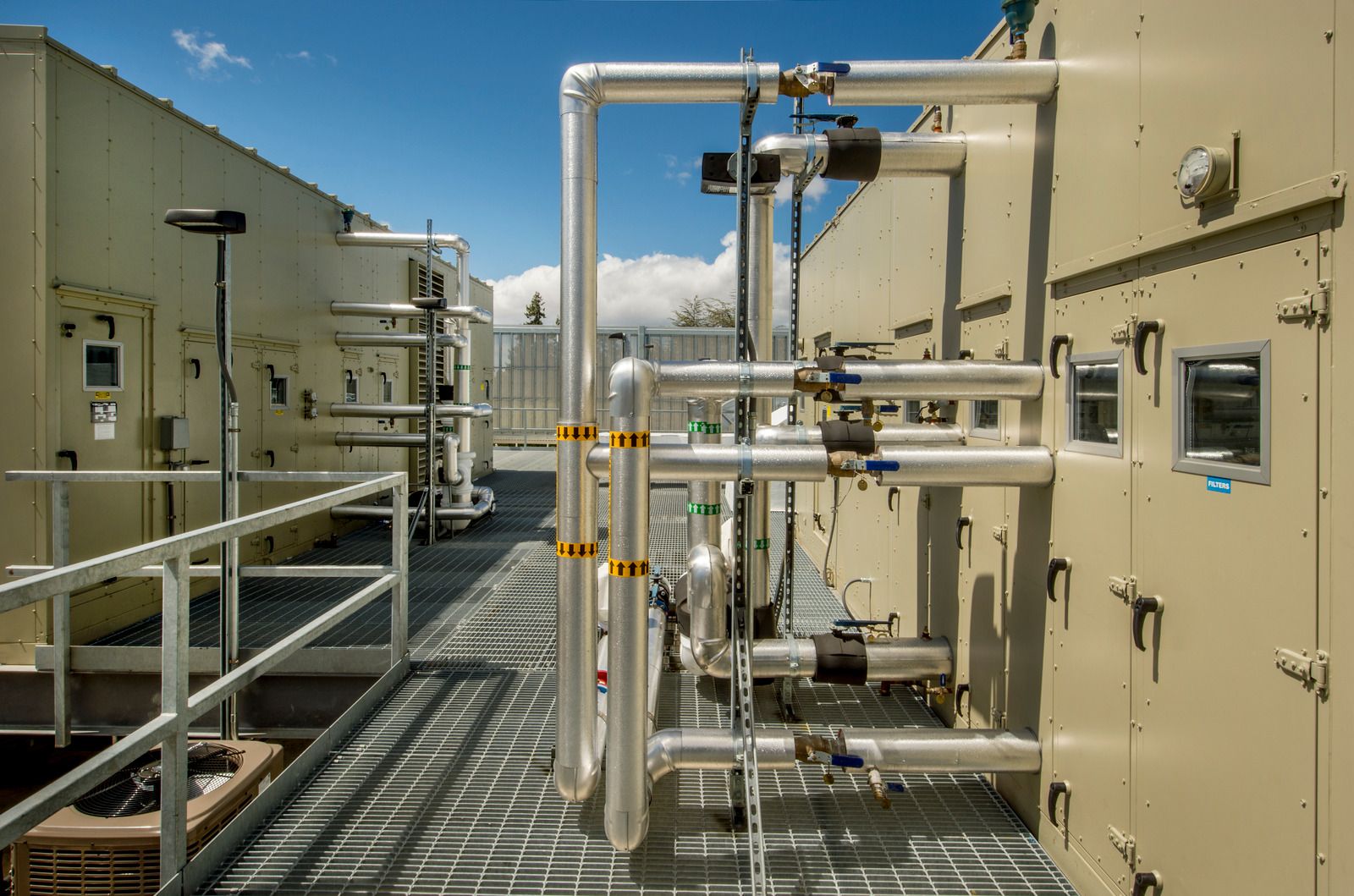 Industrial rooftop HVAC setup with insulated metal pipes, control valves, and large beige units on grated flooring under a bright blue sky, showing complex mechanical infrastructure for air handling systems.