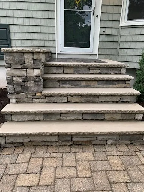 A set of stone steps with a stone-clad retaining wall lead up to a white-framed door with a dark paneled insert. The steps are constructed with light-colored concrete treads and a base of stacked stone. The entryway is framed by gray siding on the house. The ground in front of the steps is paved with interlocking rectangular stones in shades of brown and beige.