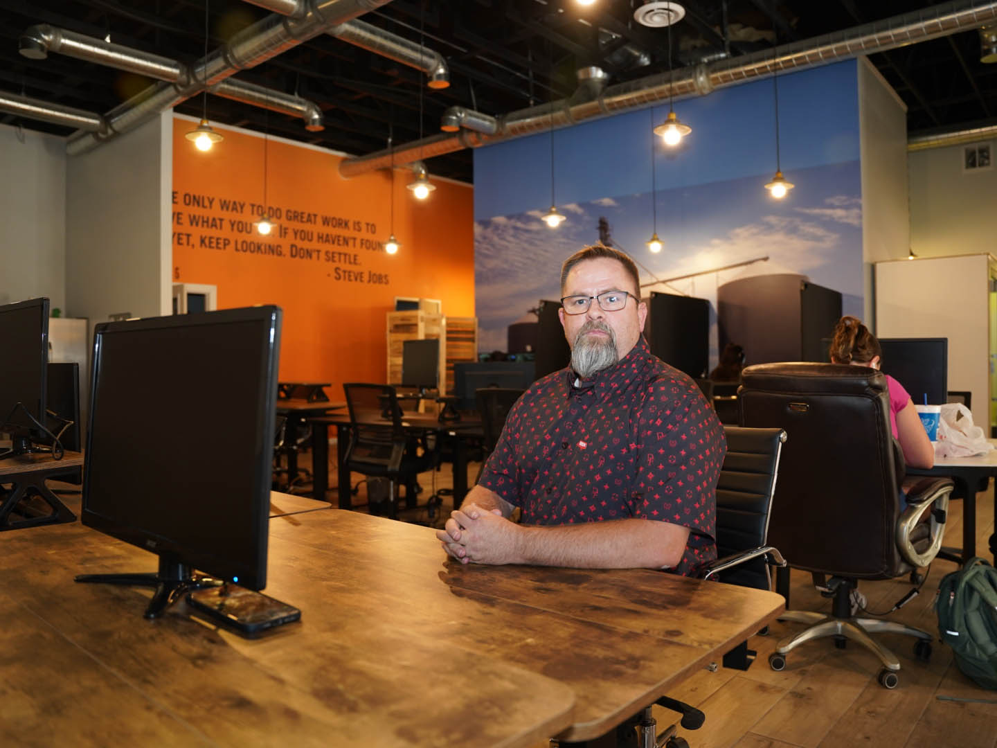 In this image, Mike Rux, an SEO professional at Ciphers Digital Marketing, is seated at a wooden desk in their Gilbert, Arizona office. He is wearing glasses and a patterned short-sleeve shirt, and his serious expression indicates focus and professionalism. The office environment includes computer monitors on wooden desks, an orange wall featuring a motivational quote by Steve Jobs, and a large mural of a scenic landscape. Other team members are seen working in the background, creating a collaborative and dynamic workspace atmosphere. The combination of motivational decor and modern office furnishings highlights the inspiring and industrious nature of the workplace.