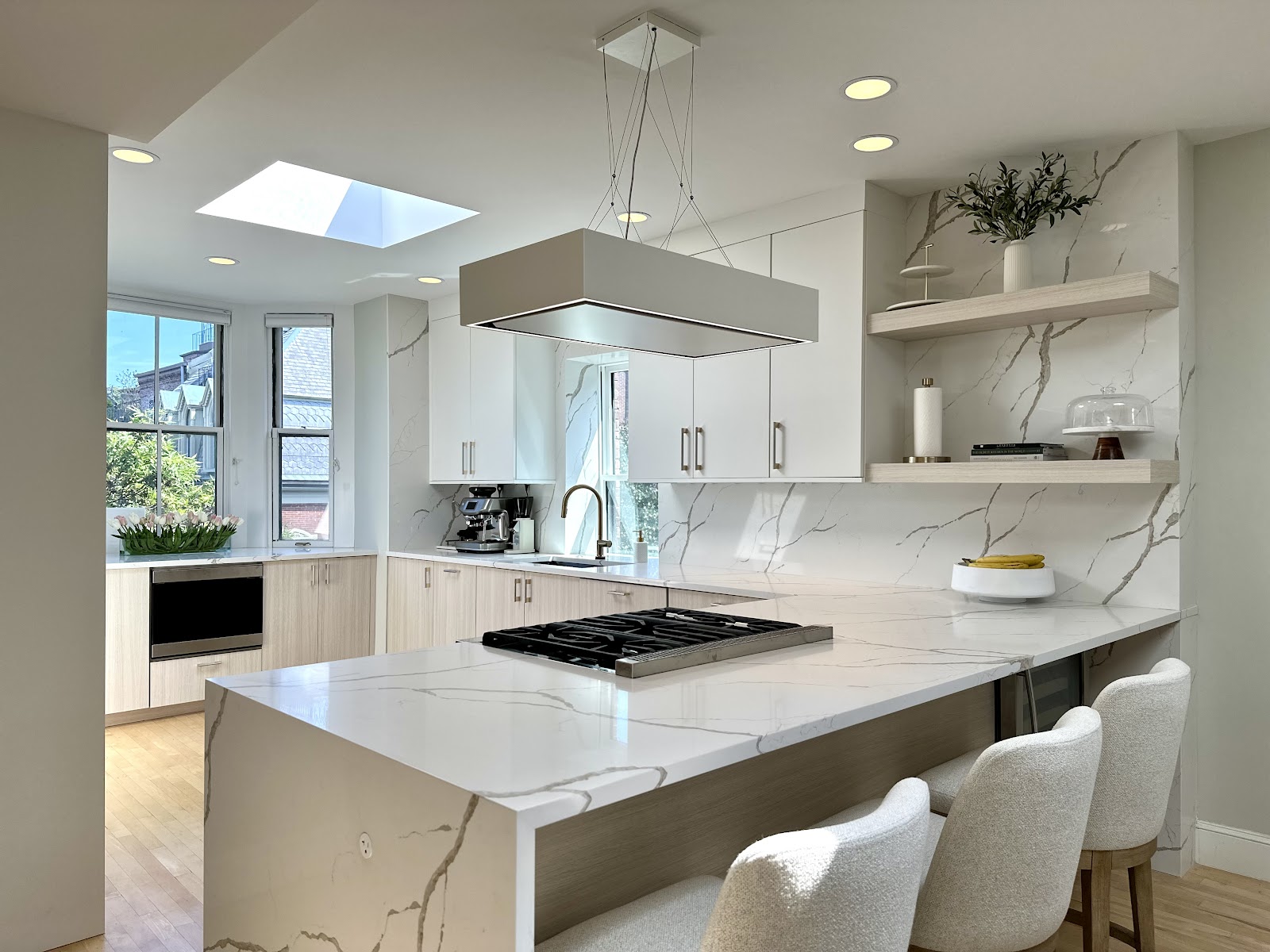 A modern, spacious kitchen with white marble countertops and light wood cabinets. A large island with a gas stovetop is in the foreground, and three plush bar stools are pulled up to the island. The kitchen features a skylight, recessed lighting, and a contemporary pendant light fixture. A coffee maker is visible on the counter, along with decorative items like a vase of flowers and a cake stand.