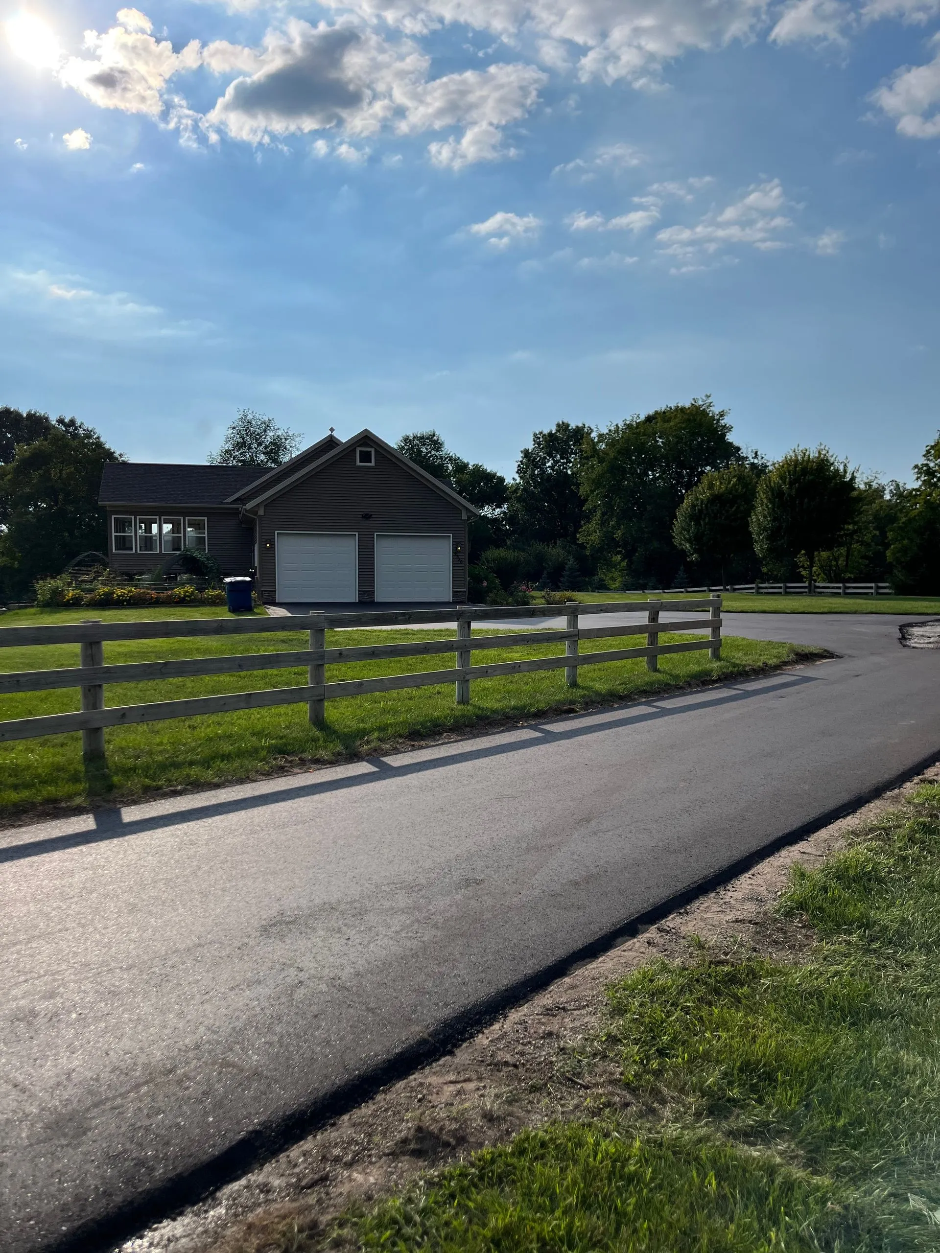 Newly completed asphalt driveway leading to a rural home with wooden fencing, green lawn, and bright sunny weather.