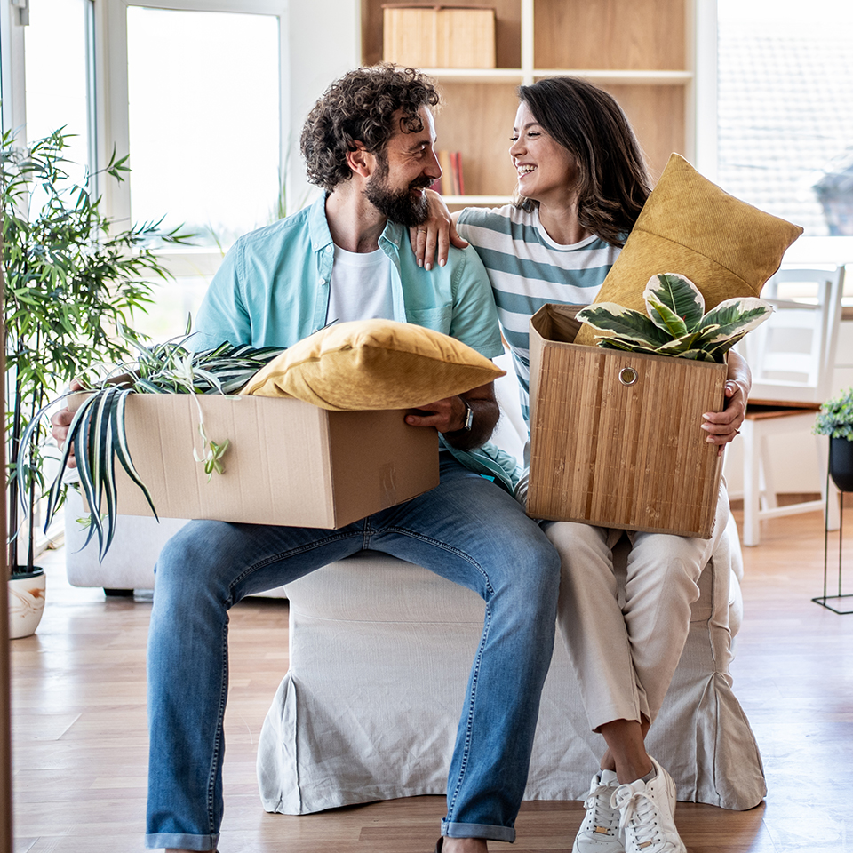 Two people holding boxes with plants and pillows inside looking at each other.