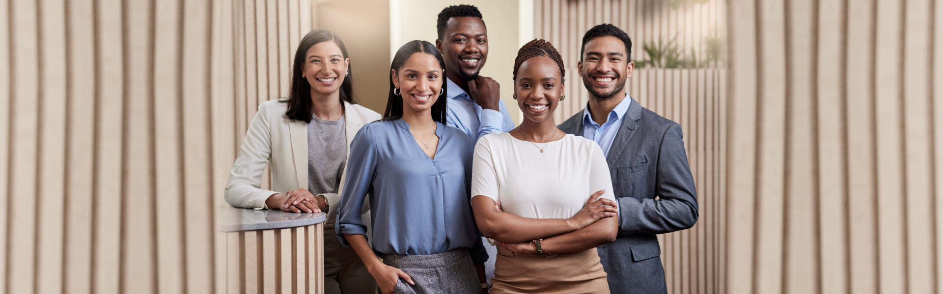 A diverse group of five business professionals, three women and two men, stand together smiling and  