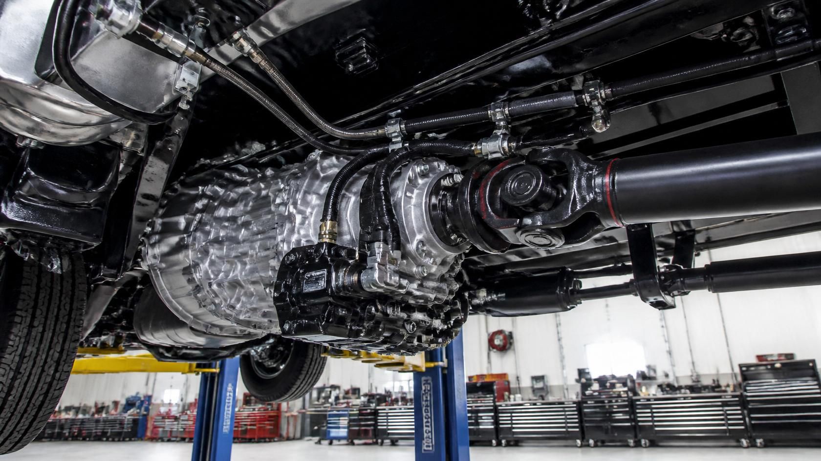 Underside view of a heavy-duty truck raised on a shop lift, showing transmission, driveshaft, universal joints, hydraulic lines, hoses, frame components, and nearby tool cabinets inside a professional repair facility.