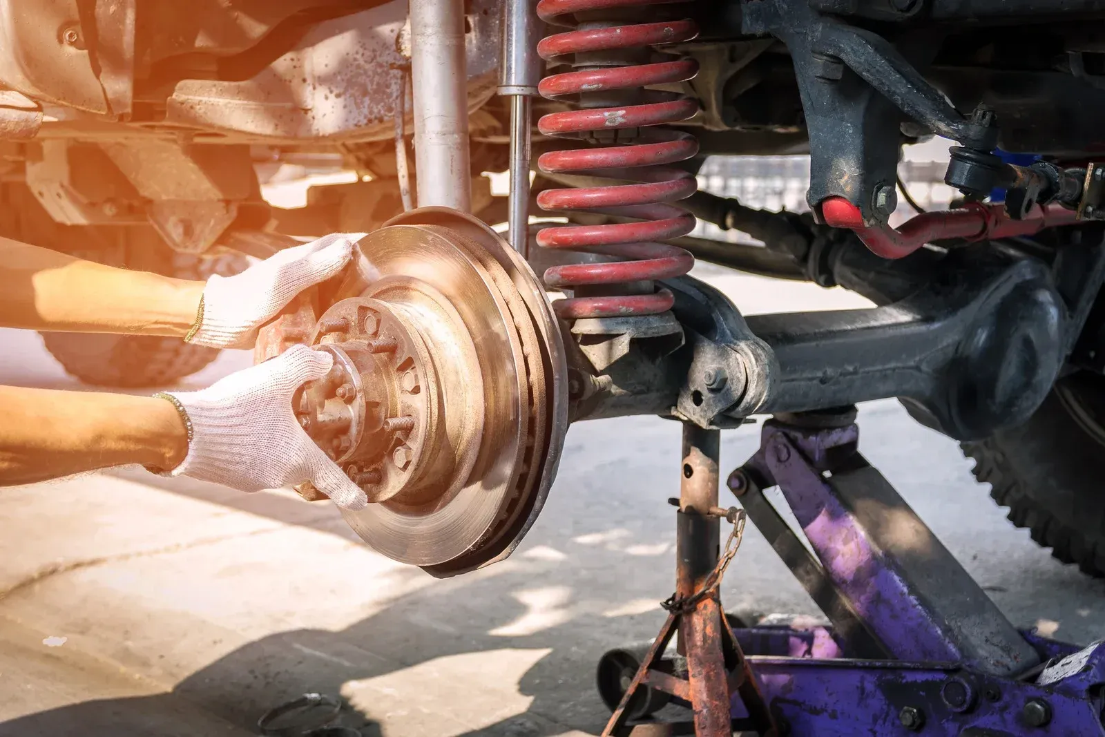 A mechanic wearing gloves installs or inspects a brake rotor on a vehicle&rsquo;s suspension system. The image highlights the brake assembly, coil spring, and shock absorber, emphasizing maintenance or repair work on automotive braking components.