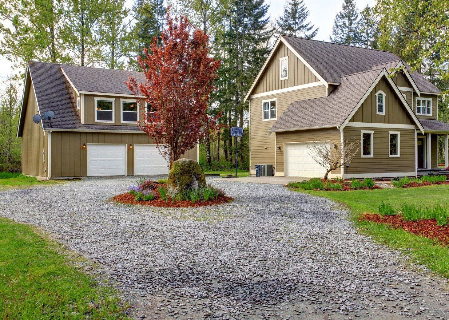 A large two-story house with a separate garage connected by a gravel driveway sits on a green lawn with mature trees under an overcast sky in a residential setting.